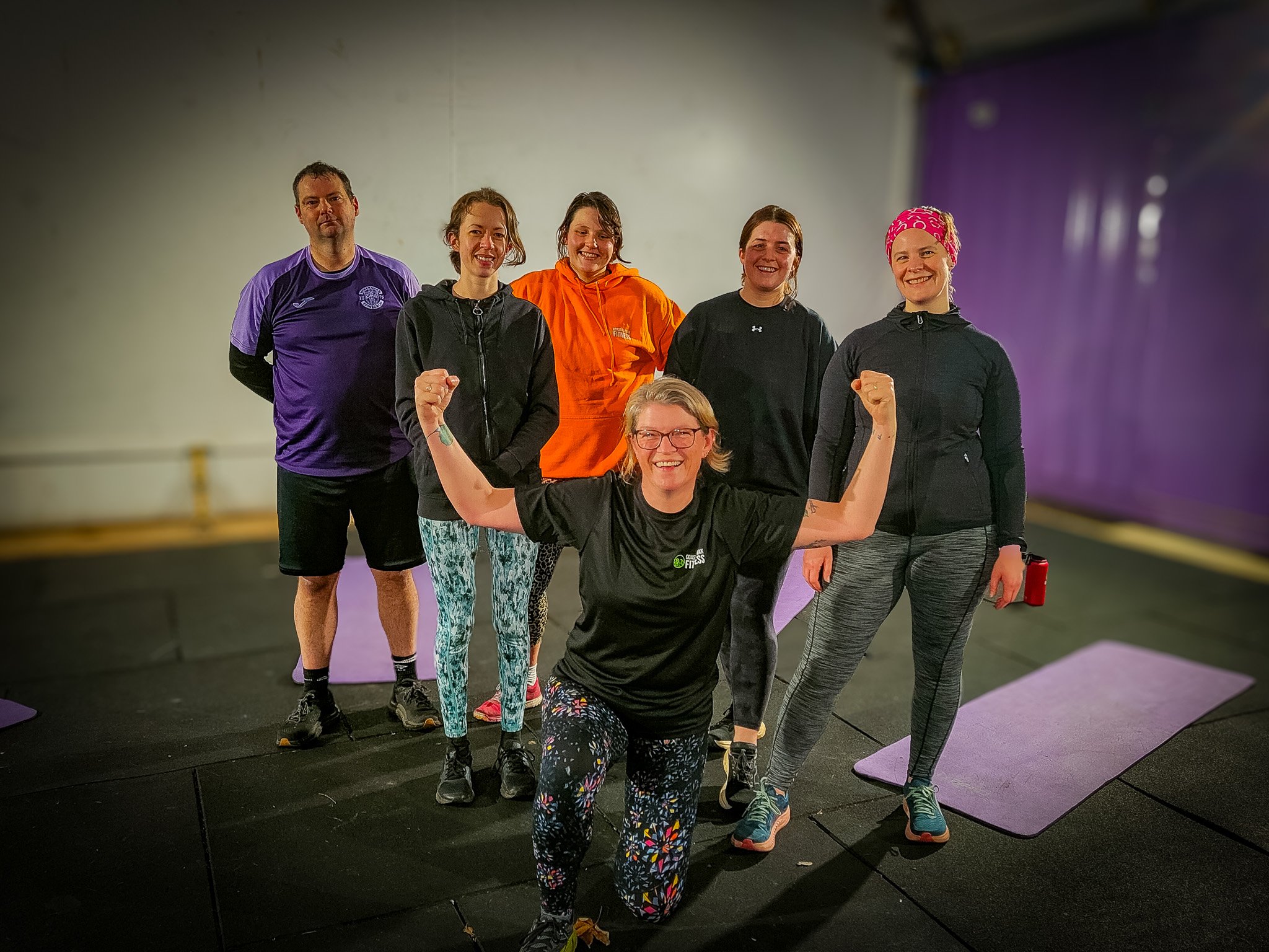 Group of six women and one man in workout attire posing in a gym, with the woman in front flexing her arms.