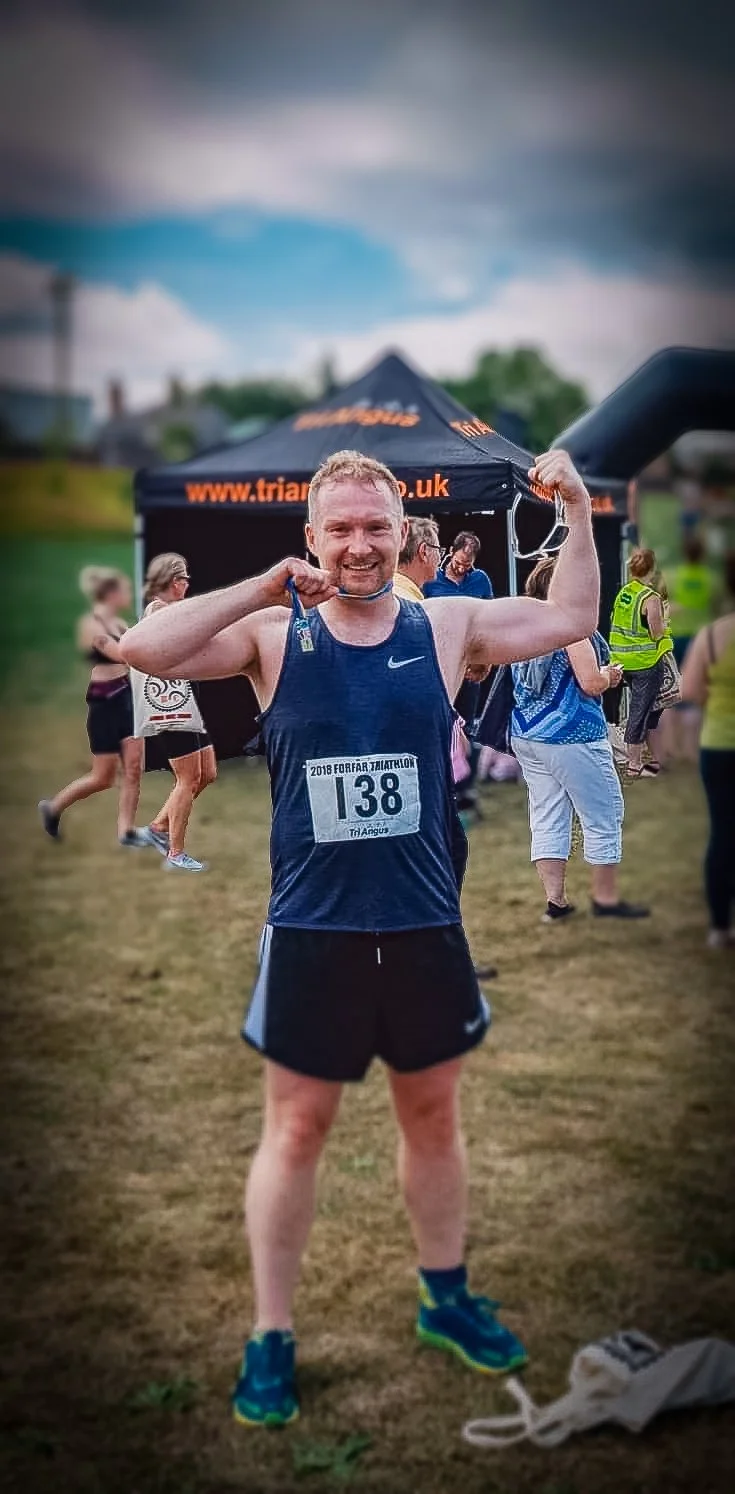 A man in athletic gear holding a medal at a triathlon event, smiling, with other participants and event tents in the background.