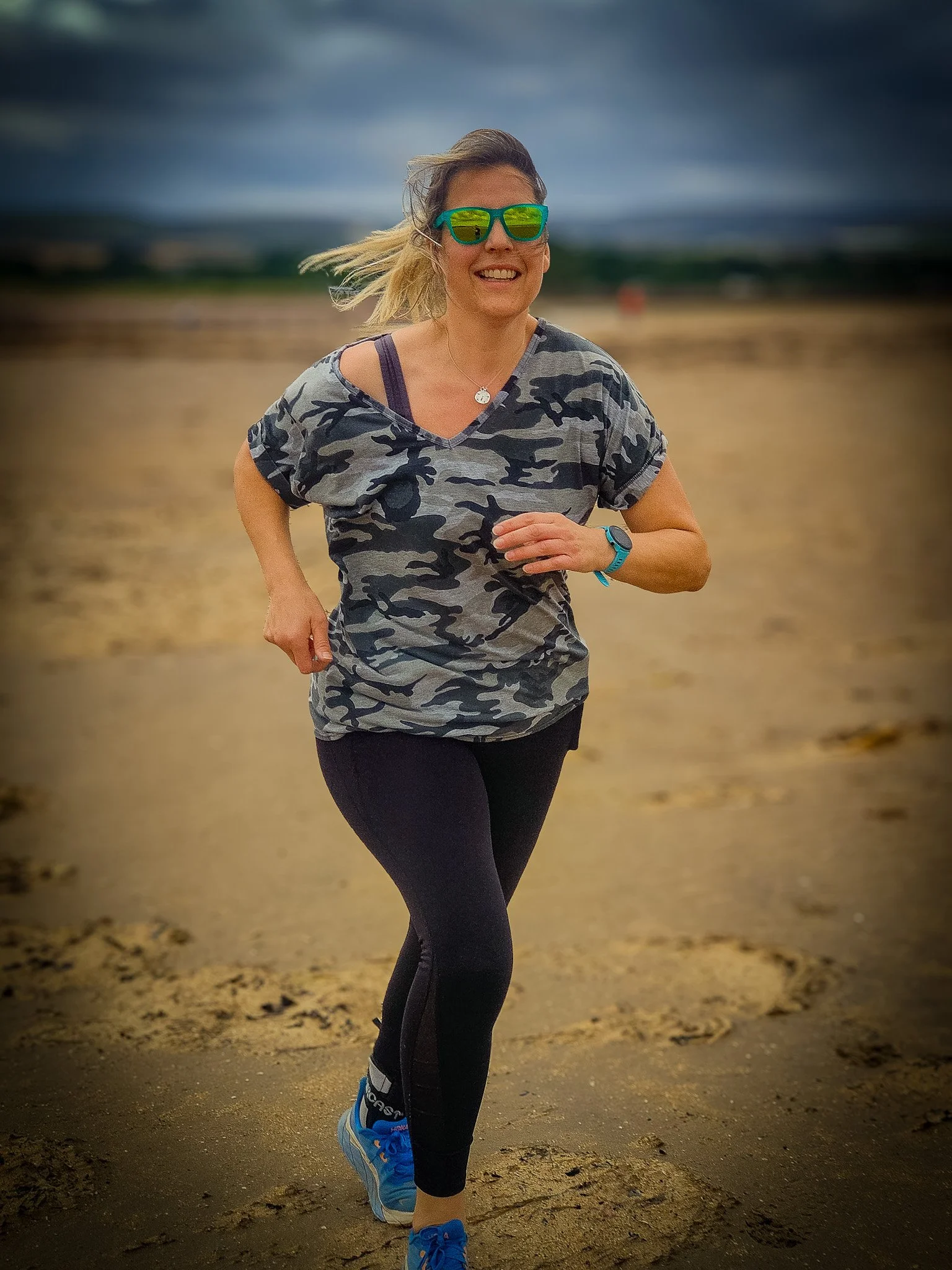 Woman running on a sandy beach with a cloudy sky, wearing sunglasses, a camouflage t-shirt, black leggings, and blue running shoes.