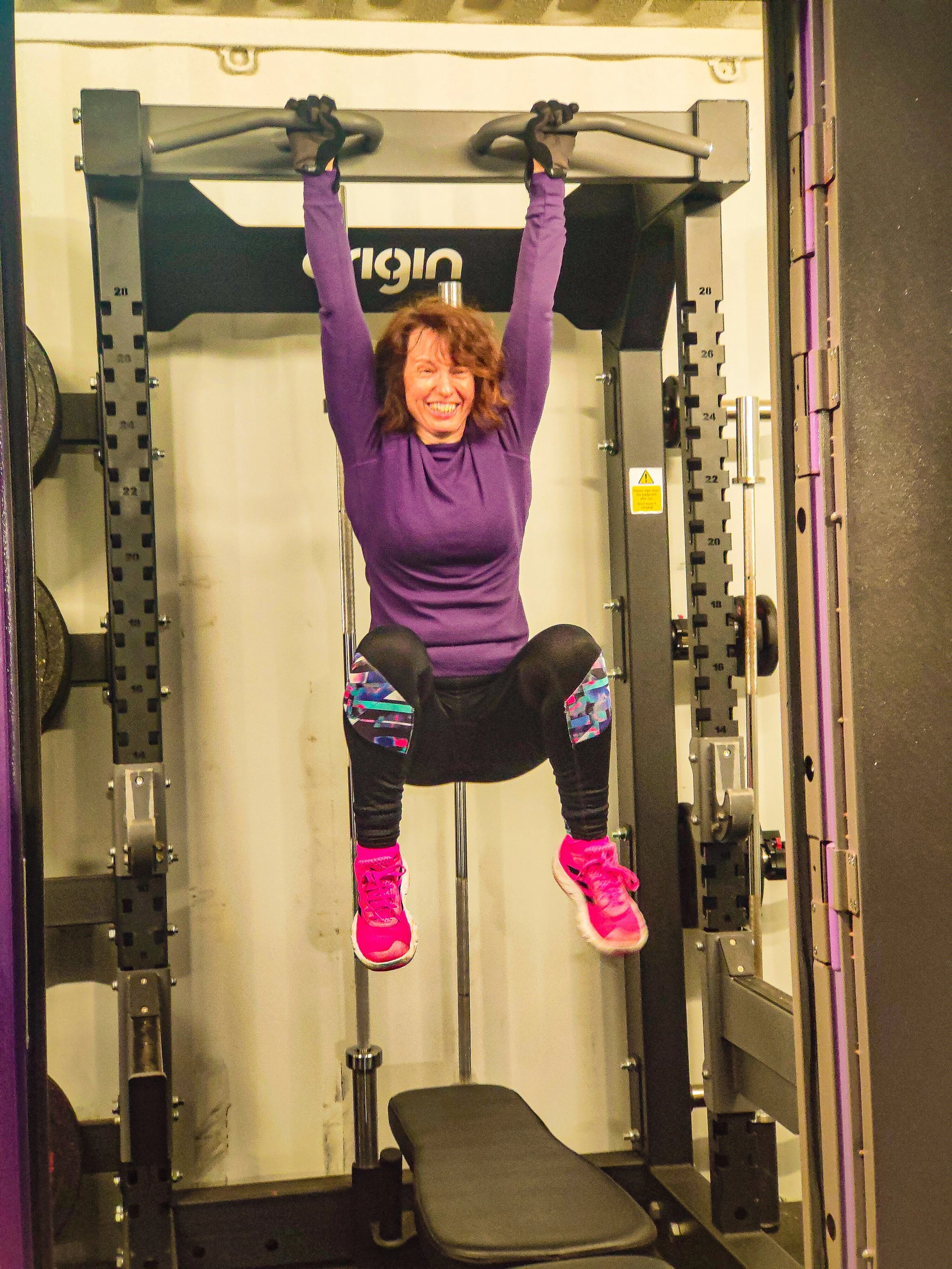 A woman hanging from a pull-up bar in a gym, smiling and wearing a purple long-sleeve shirt, black leggings with colorful patterns, and pink sneakers.