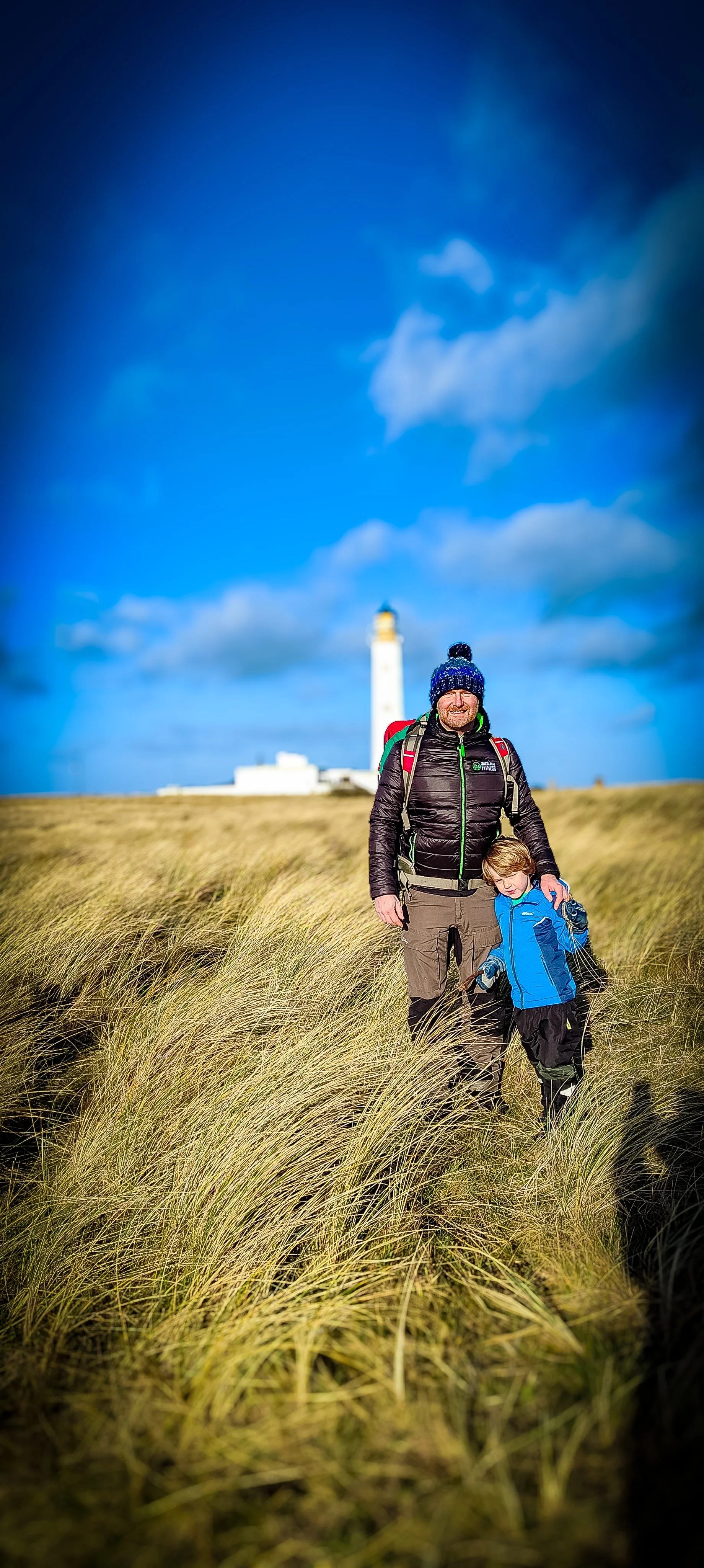 A man and a young boy hiking through tall grass near a lighthouse on a cloudy day.