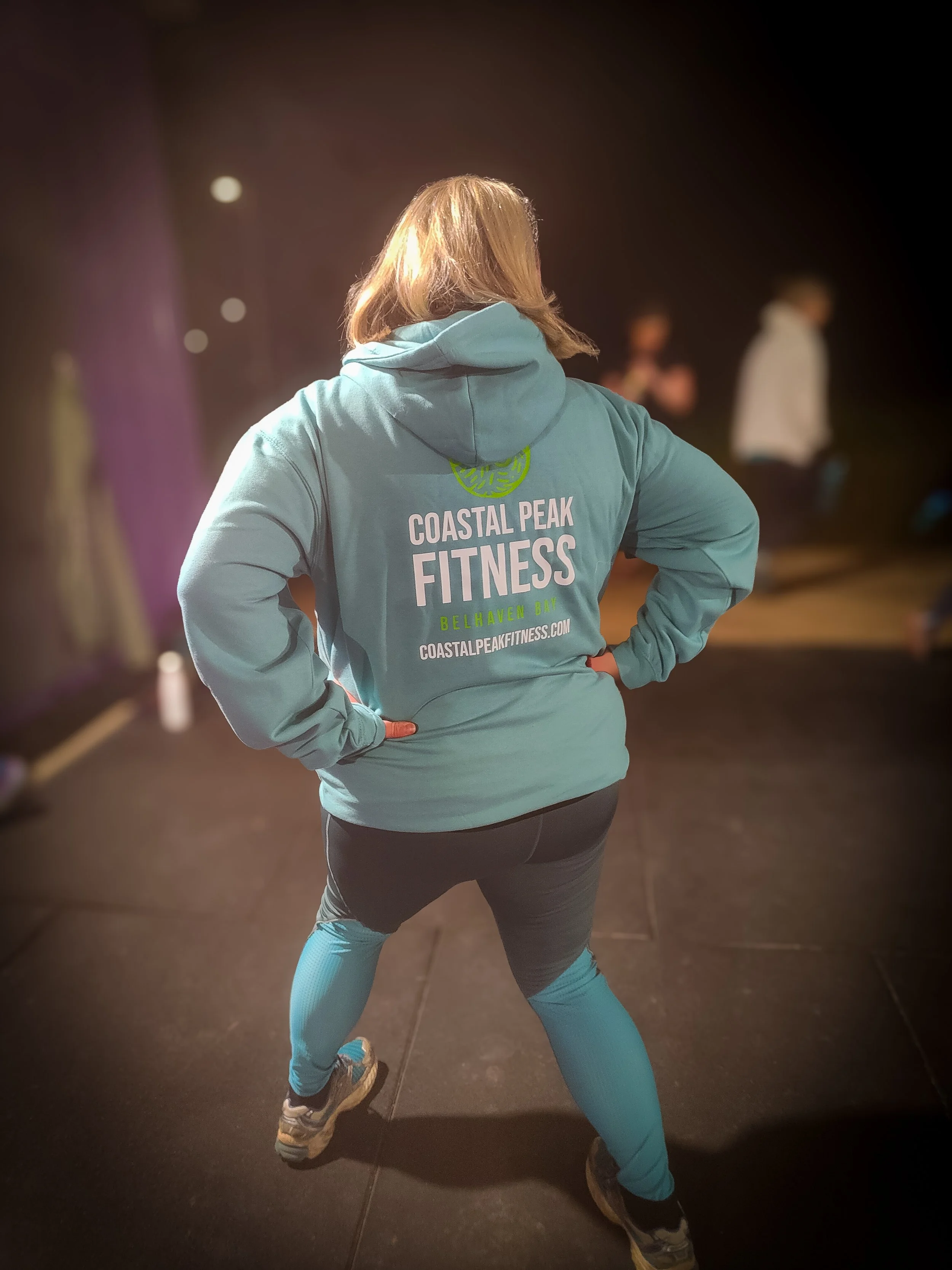 Back view of a woman in athletic wear, standing with hands on hips, facing away, in a gym or fitness studio, wearing a hoodie with 'Coastal Peak Fitness' logo on the back.