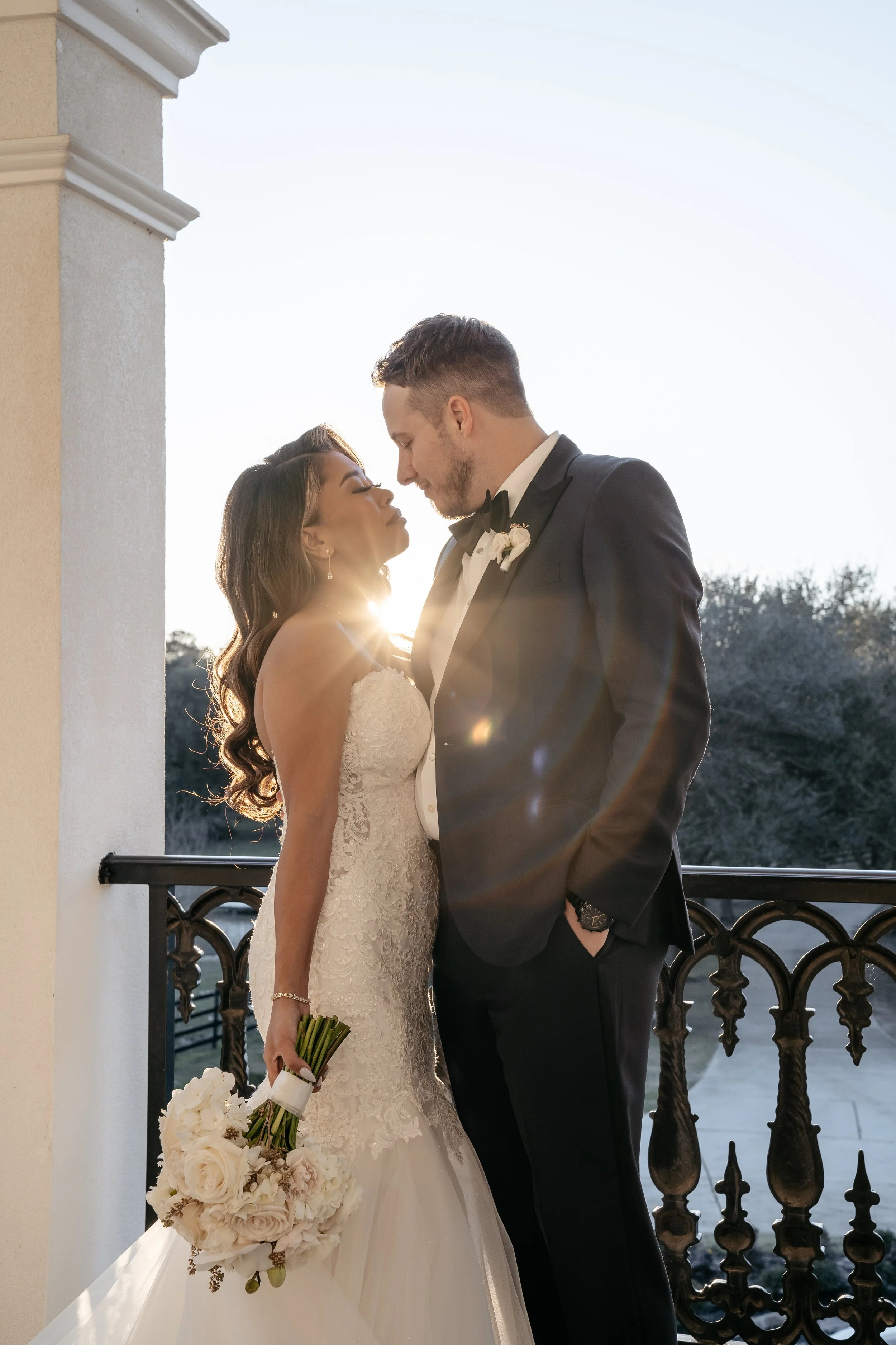 A bride and groom standing close together on a balcony during sunset, with the bride holding a bouquet of white flowers and the sun creating a lens flare.