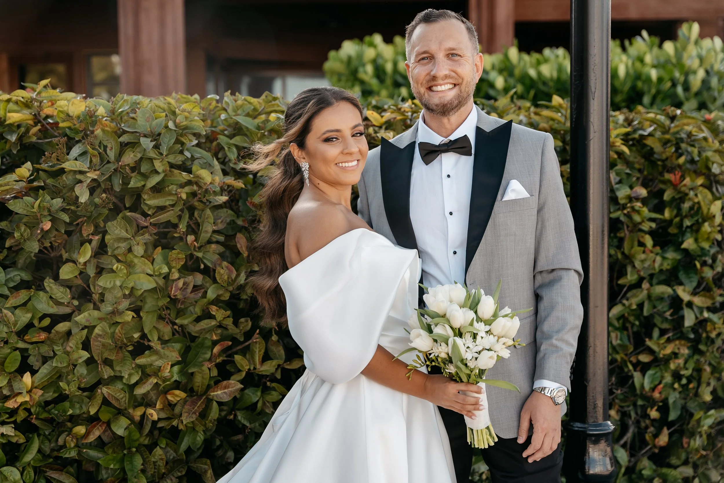 A smiling bride in a white wedding dress holding a bouquet of white tulips and a groom in a gray tuxedo with a black bow tie, standing outdoors in front of green bushes.