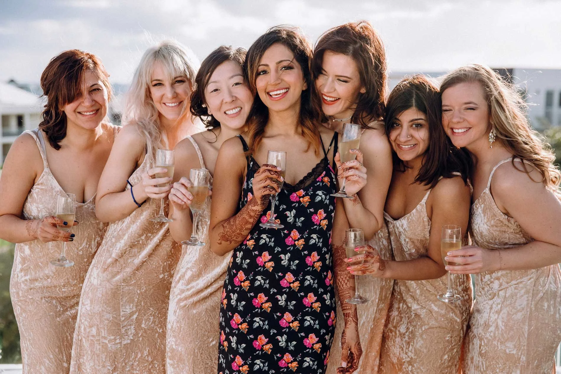 A group of women in floral and light-colored dresses celebrating with champagne outdoors on a sunny day.