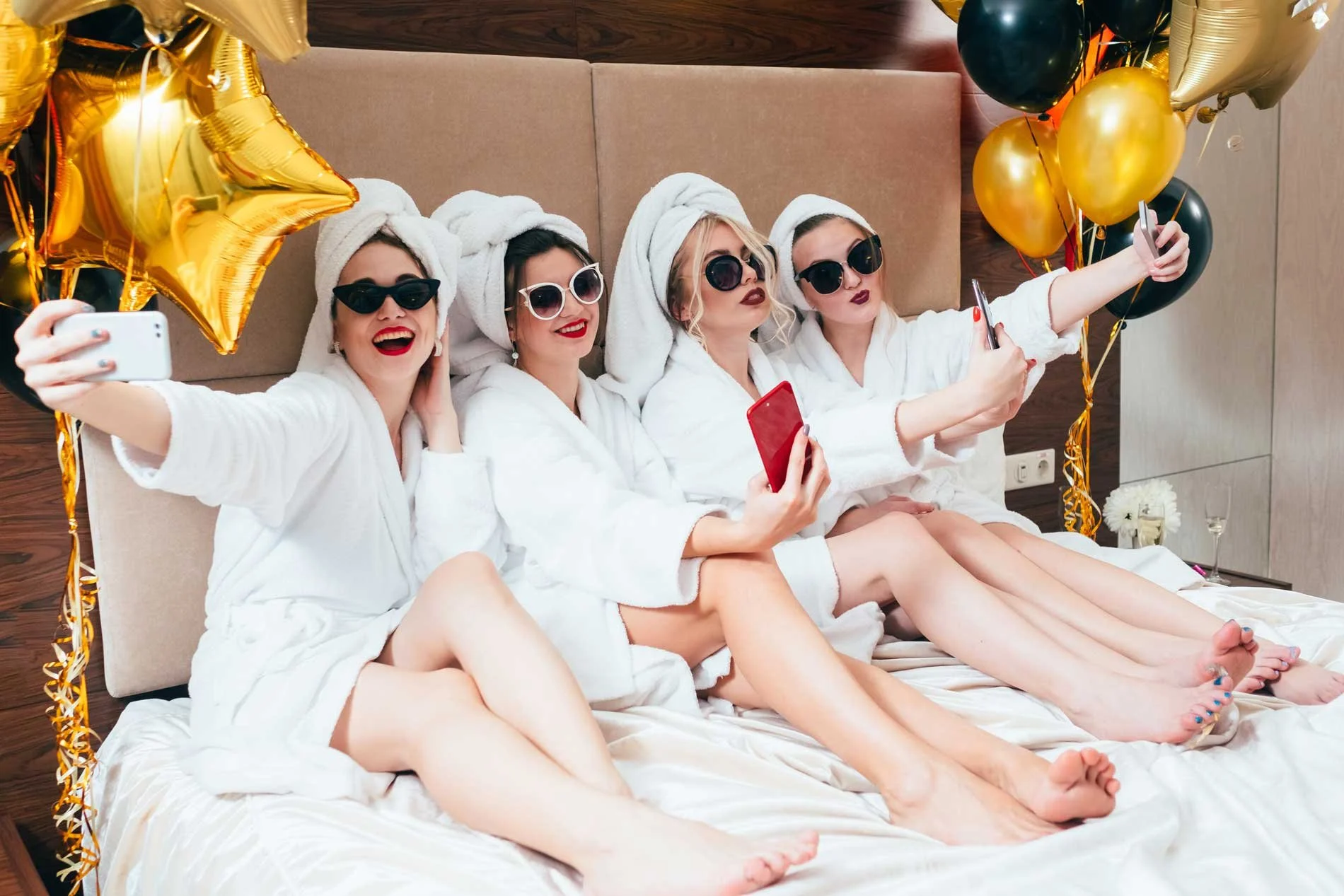Four women in bathrobes with towels on heads, wearing sunglasses, sitting on a bed with balloons around them, taking selfies and smiling.