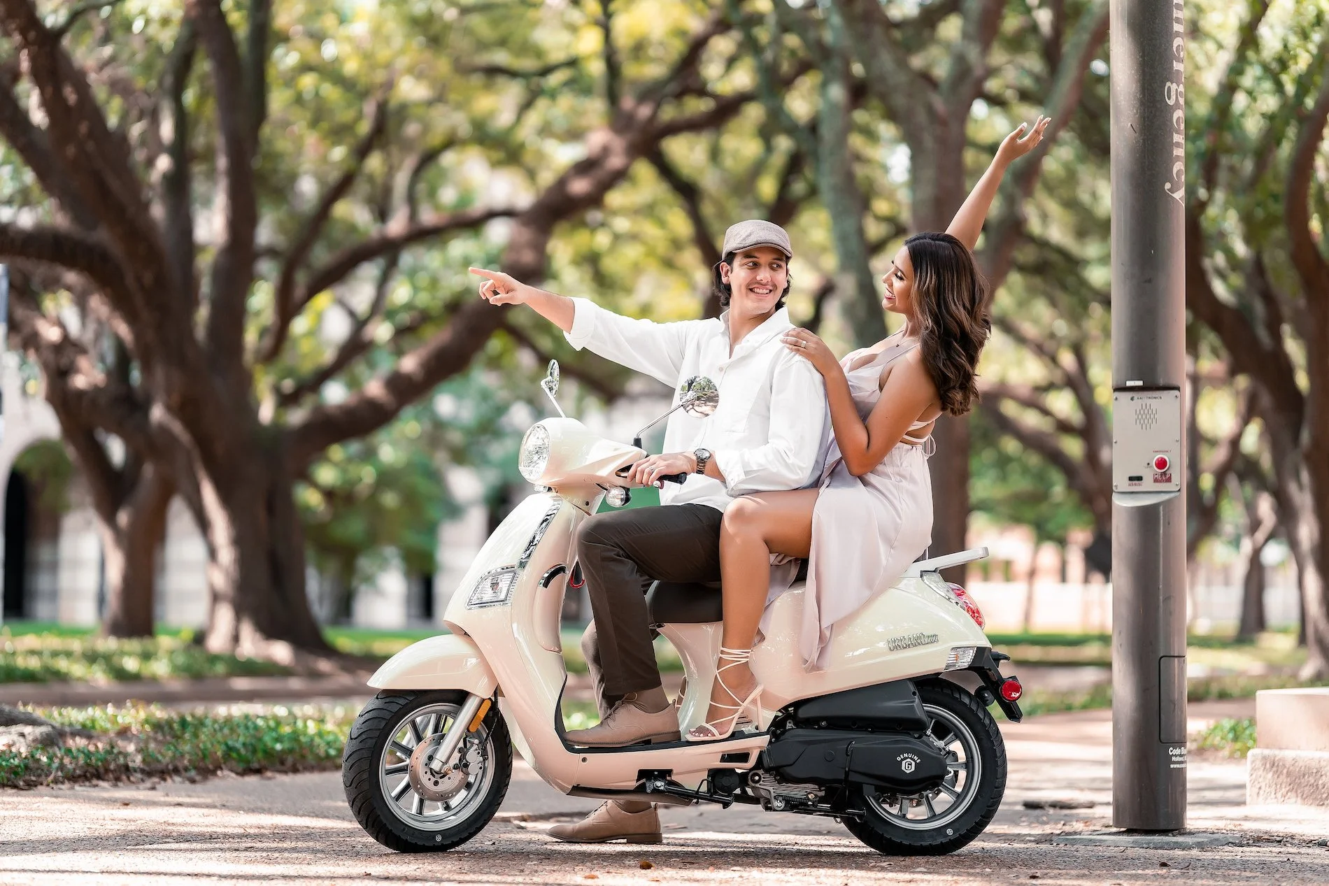 A couple riding a scooter in a park with trees, the woman sitting behind the man and holding onto him, both smiling and enjoying a sunny day.