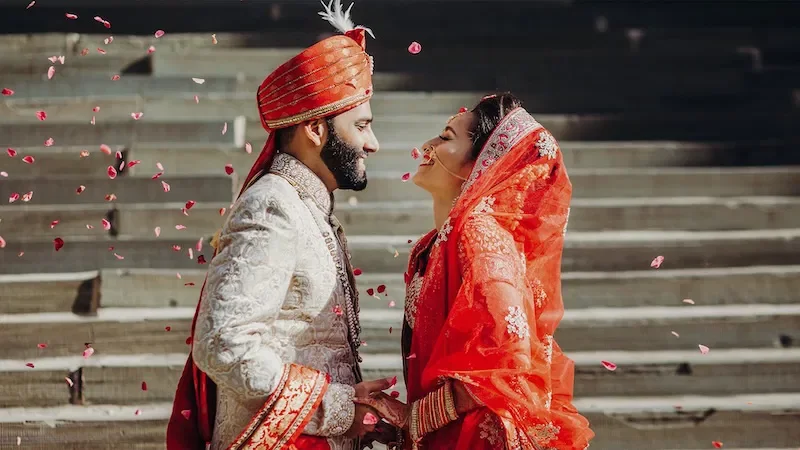 An Indian wedding couple smiling and facing each other, surrounded by falling rose petals, with wooden stairs in the background.