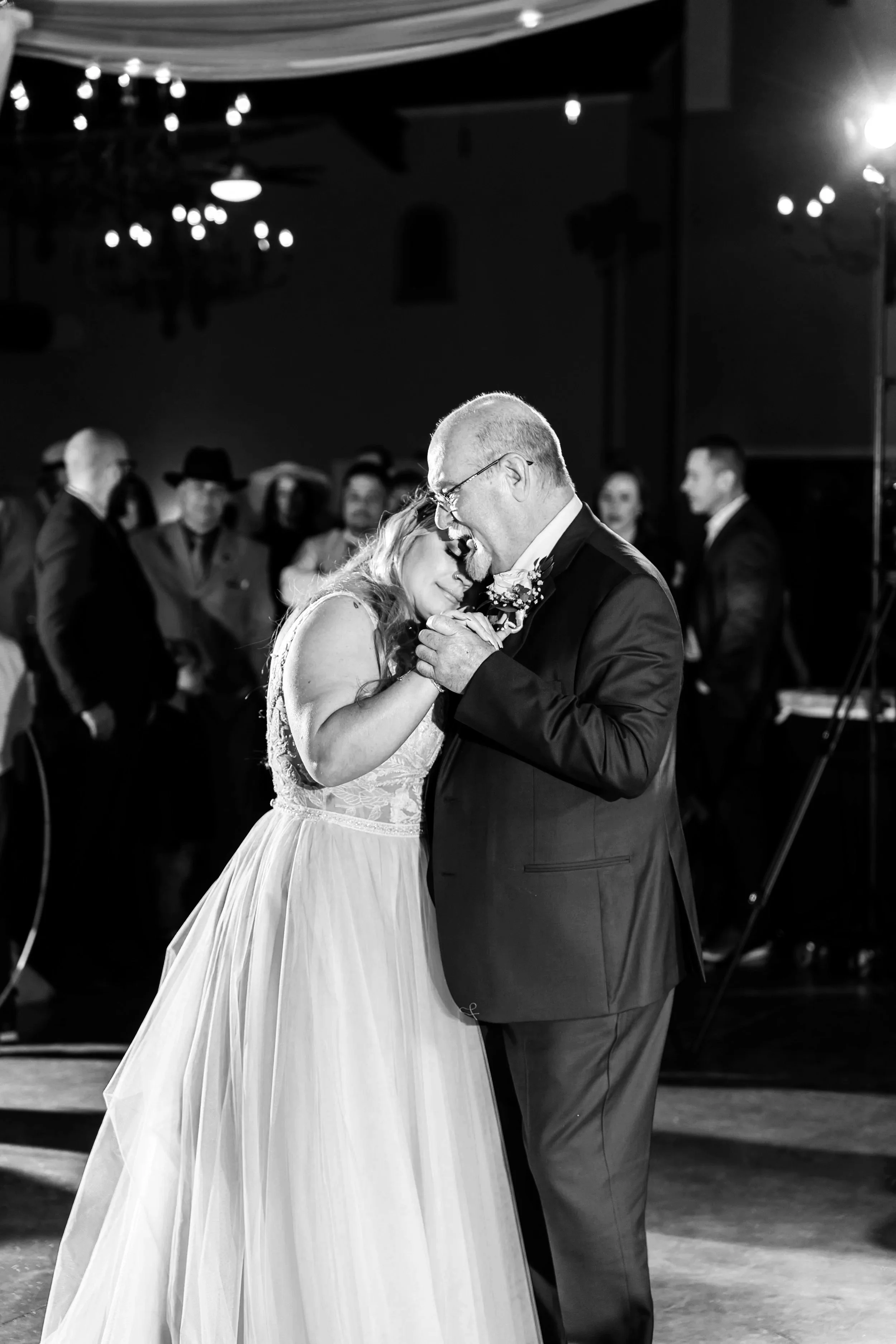 A bride and an older man, possibly her father, share a dance at a wedding reception. The bride’s head rests on the man’s chest as they hold hands. The background features several guests watching the dance.