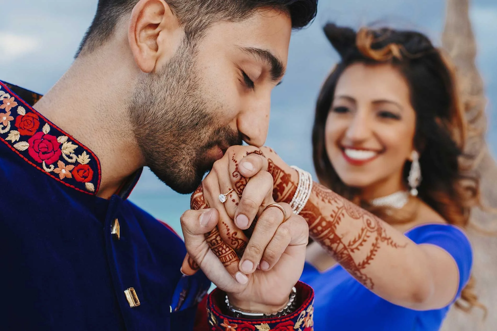 A man in a navy blue traditional outfit with floral embroidery is kissing a woman's hand, who is smiling in the background. The woman is dressed in a blue dress with jewelry and henna on her hand.