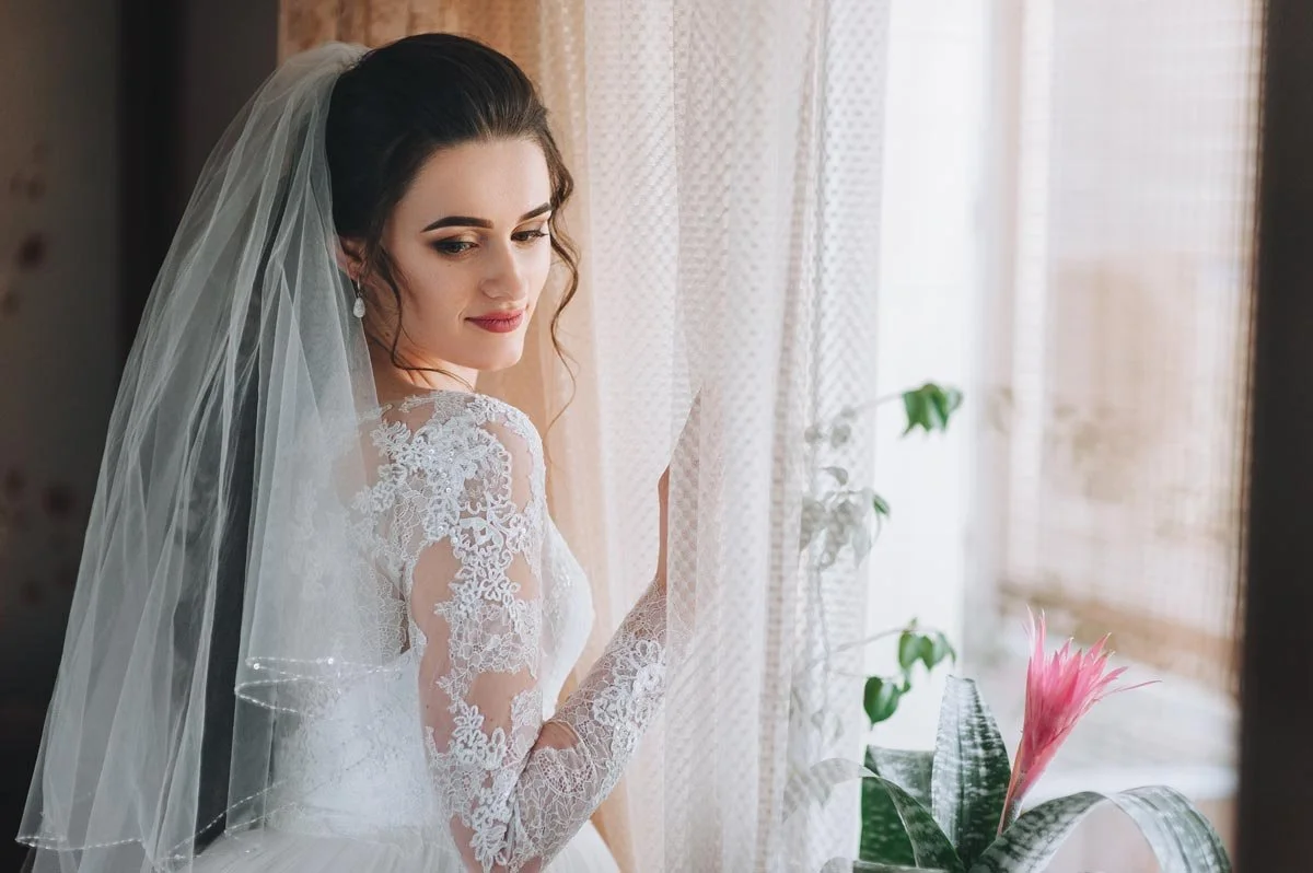 Bride in a lace wedding gown and veil standing by a window, captured in a soft, romantic bridal portrait
