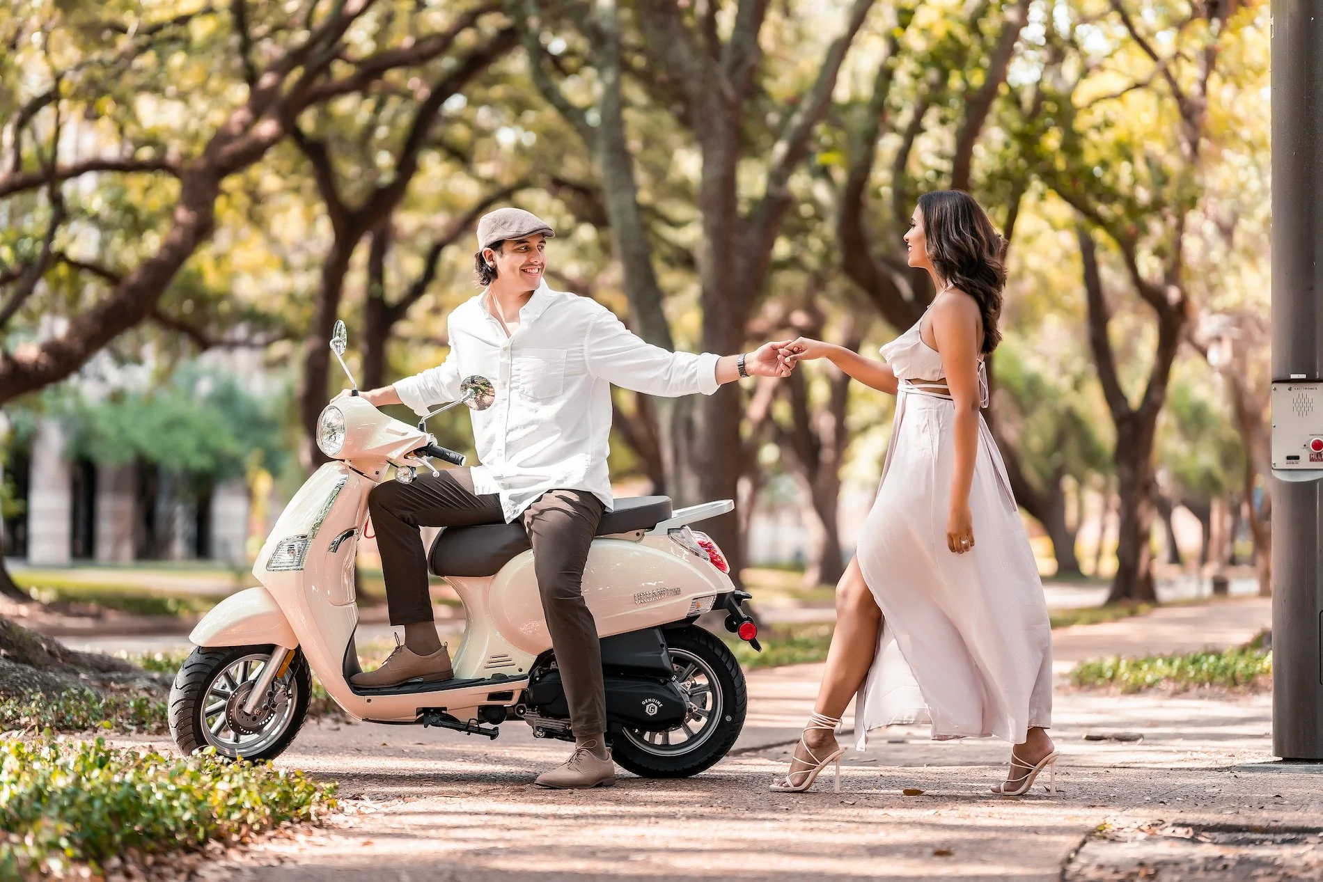 A man on a pink scooter handing a woman part of his ring while standing on a sidewalk in a park with trees in the background.
