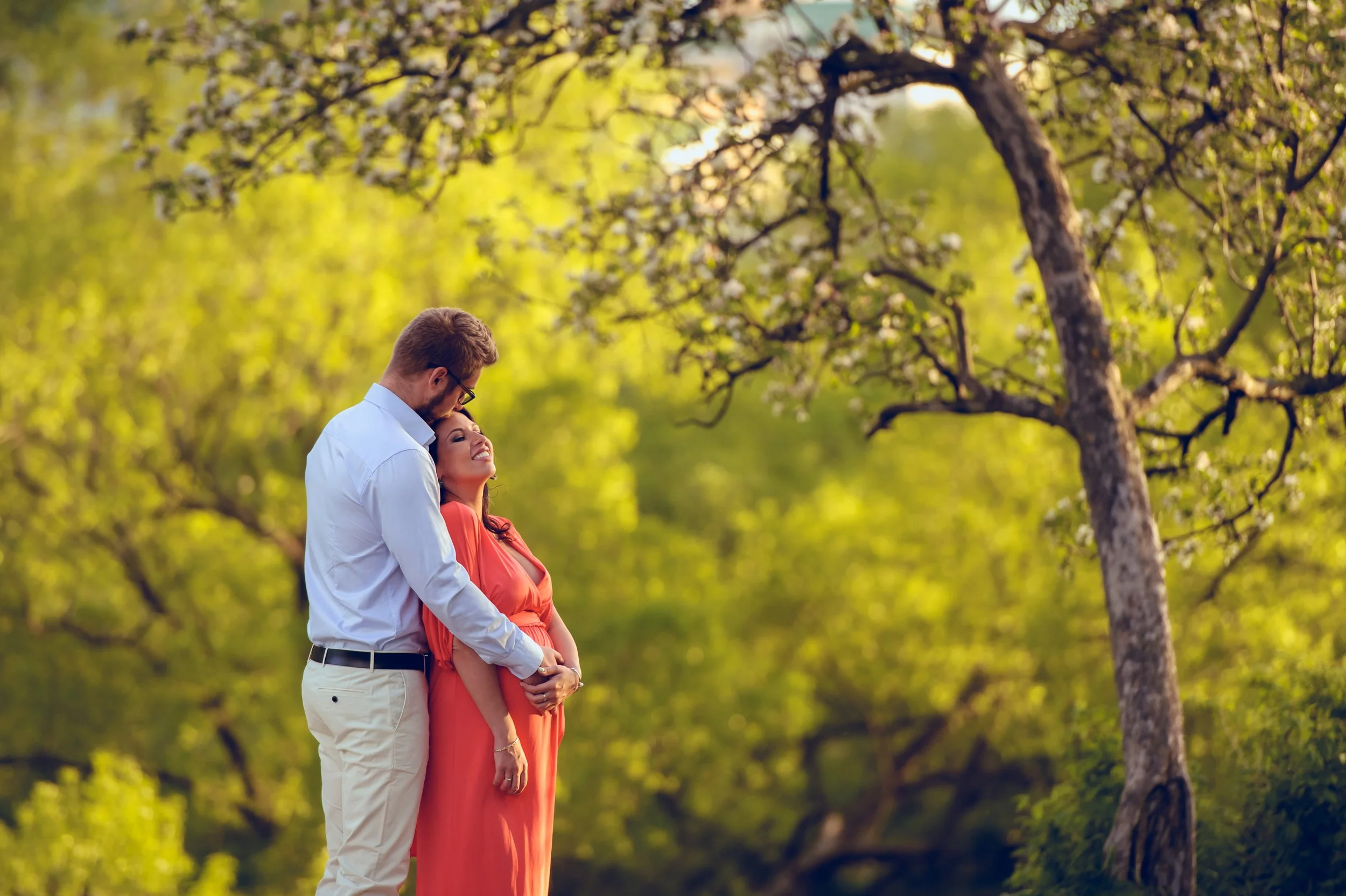 A couple, a man in a white shirt and a woman in an orange dress, standing close outdoors under a tree, smiling and enjoying each other's company with a lush green background.