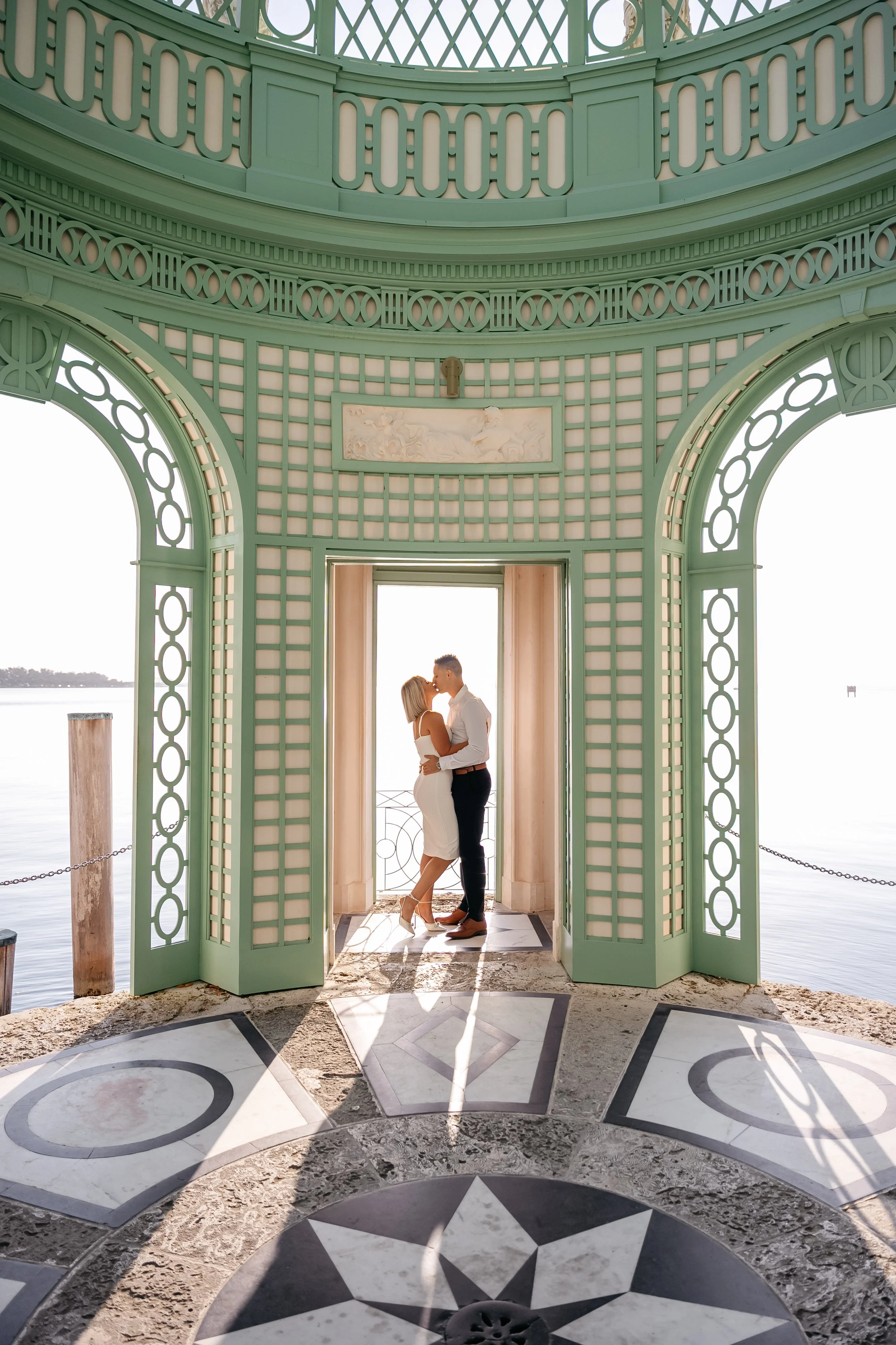 A couple kissing in an ornate, green, open-air pavilion overlooking water, with patterned marble flooring.