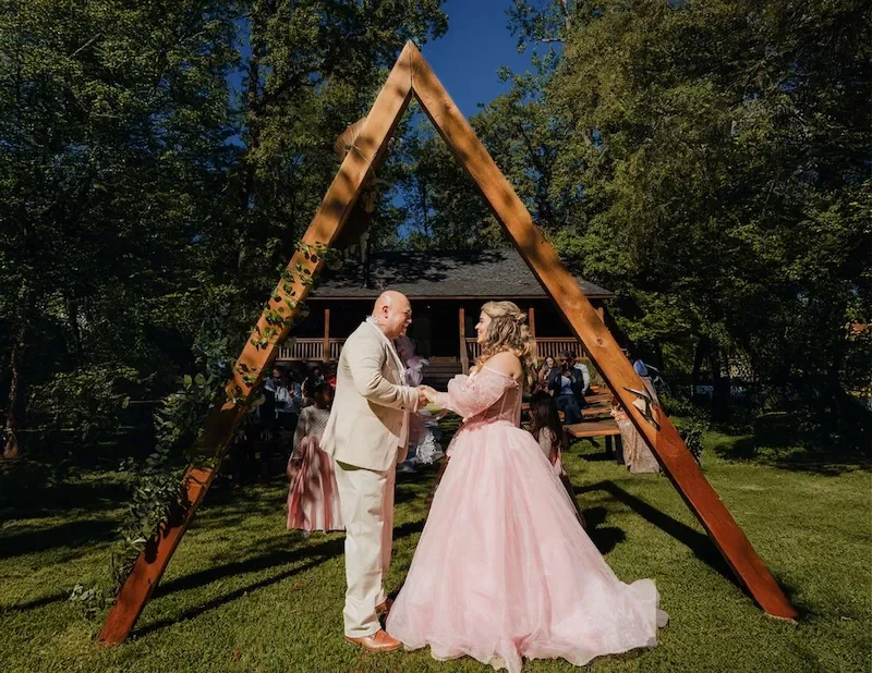 A couple getting married outdoors beneath a wooden A-frame arch decorated with green foliage, with guests seated behind them on a grassy lawn and a house in the background.