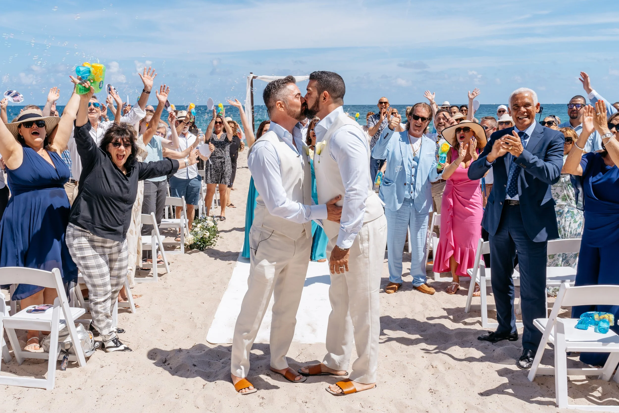 Two men kissing at a beach wedding ceremony while surrounded by celebrating guests, with the ocean in the background.