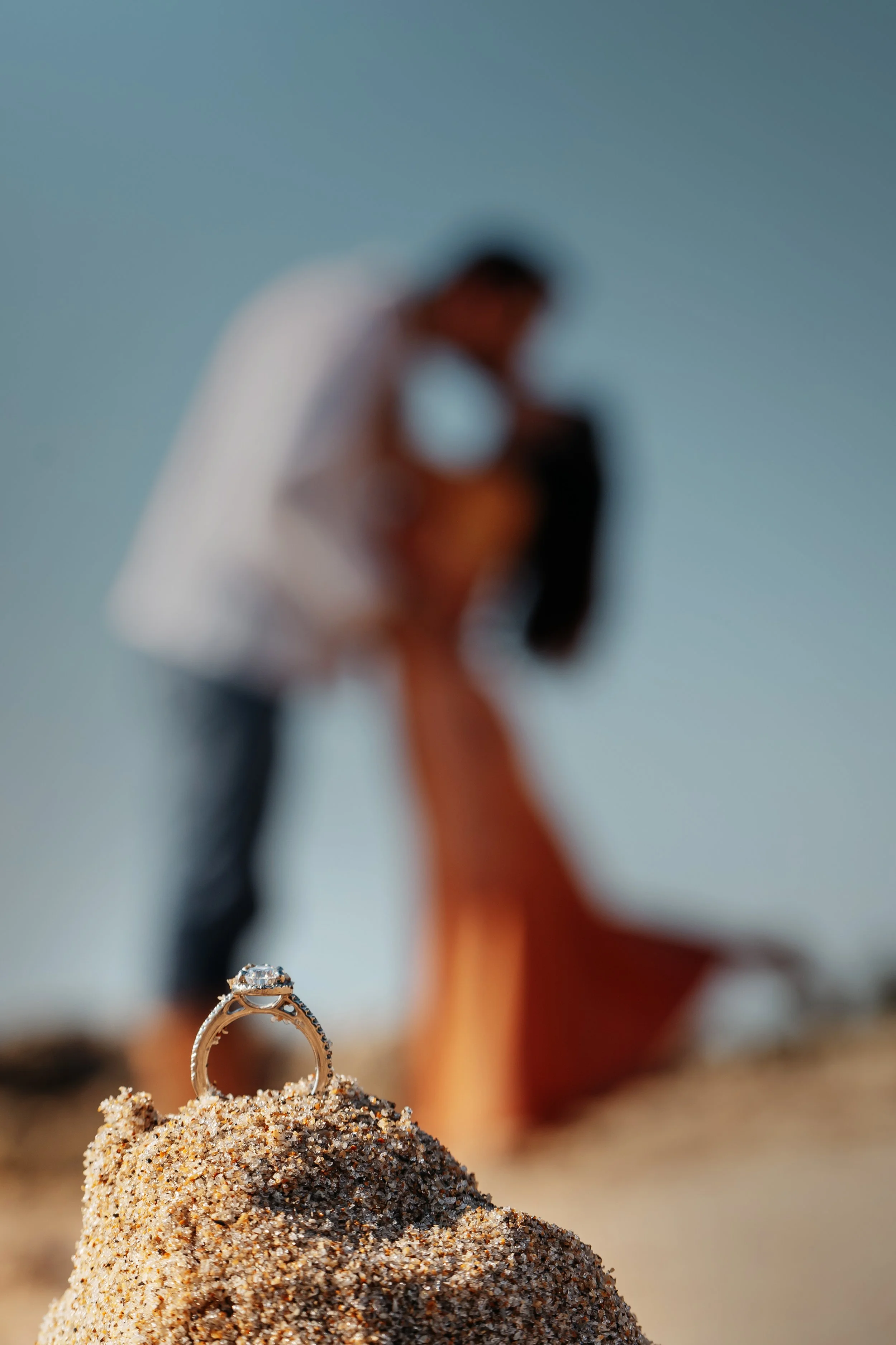 A close-up of a diamond engagement ring on a mound of sand with a blurred background of a couple on the beach, where the man is kneeling and the woman is sitting.