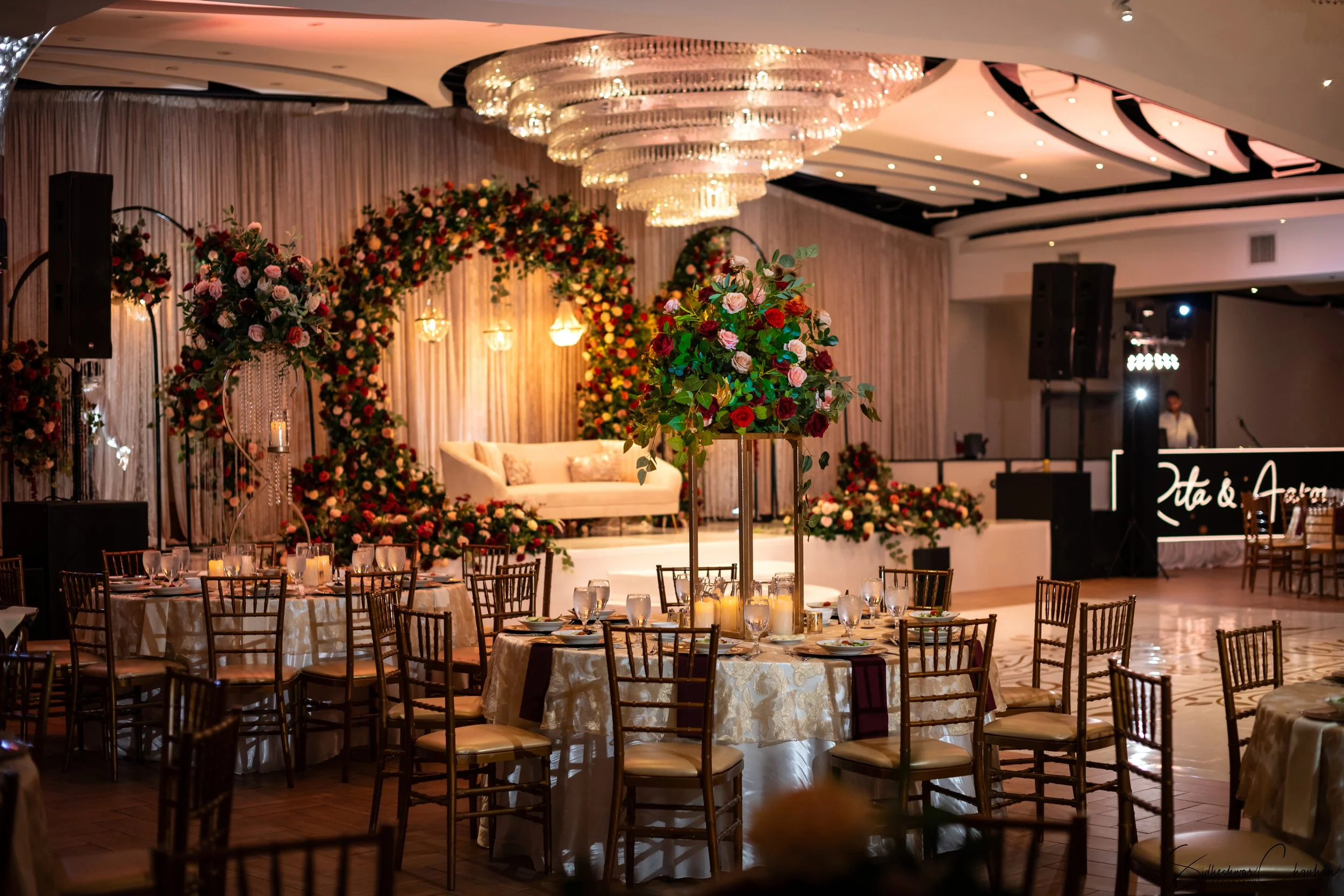 Elegant wedding reception hall with round tables decorated with floral centerpieces, a stage with a white sofa and floral arches, and chandeliers hanging from the ceiling.