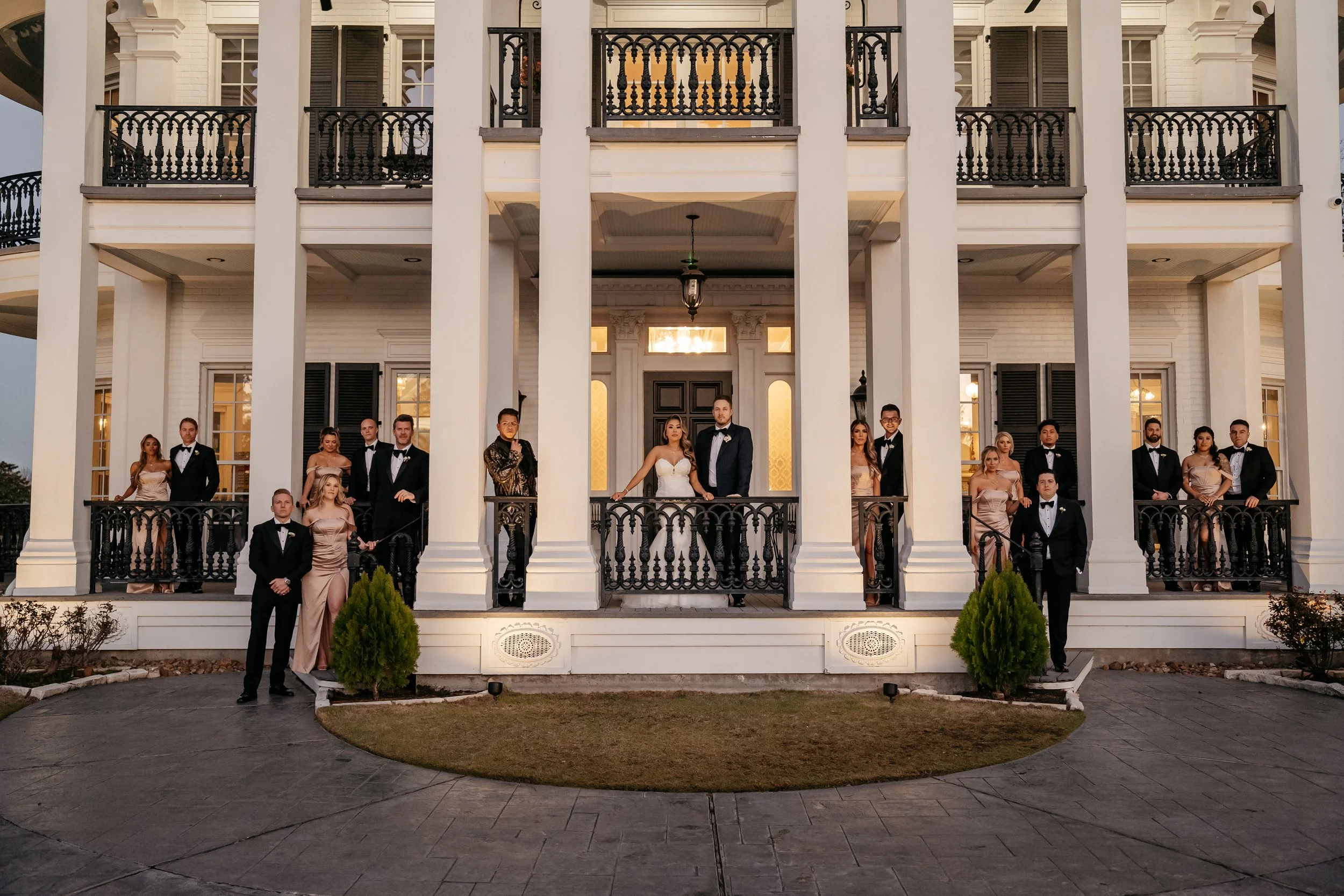 A wedding party standing on the front steps and balconies of a large, white, multi-story house with black railings and windows. The bride and groom are at the center, with the bride in a white wedding dress and the groom in a dark tuxedo. Bridesmaids