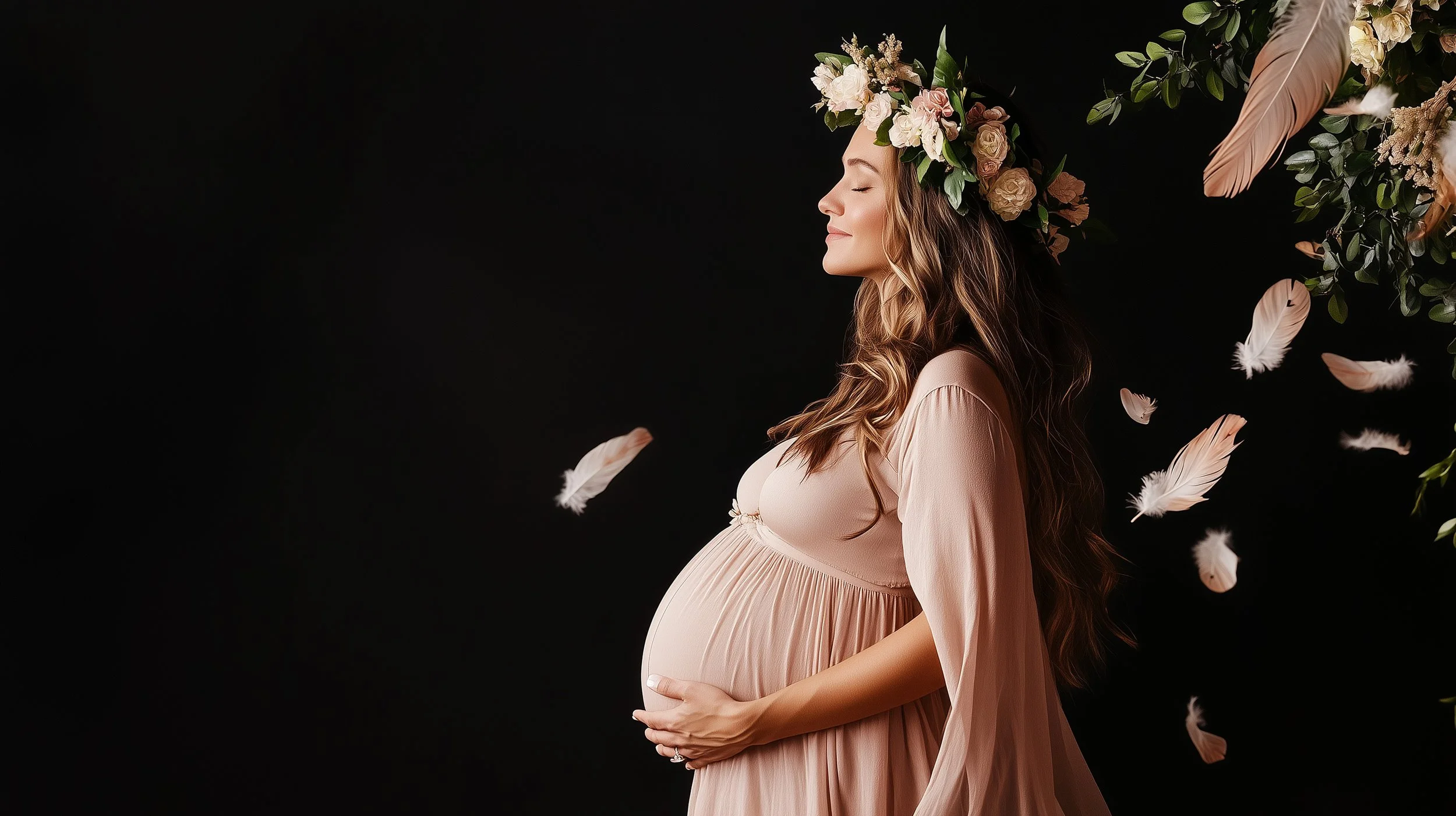 A pregnant woman in a beige dress with a floral crown on her head, standing sideways with her hands on her belly, and closed eyes. There are soft pink feathers and green foliage around her against a black background.