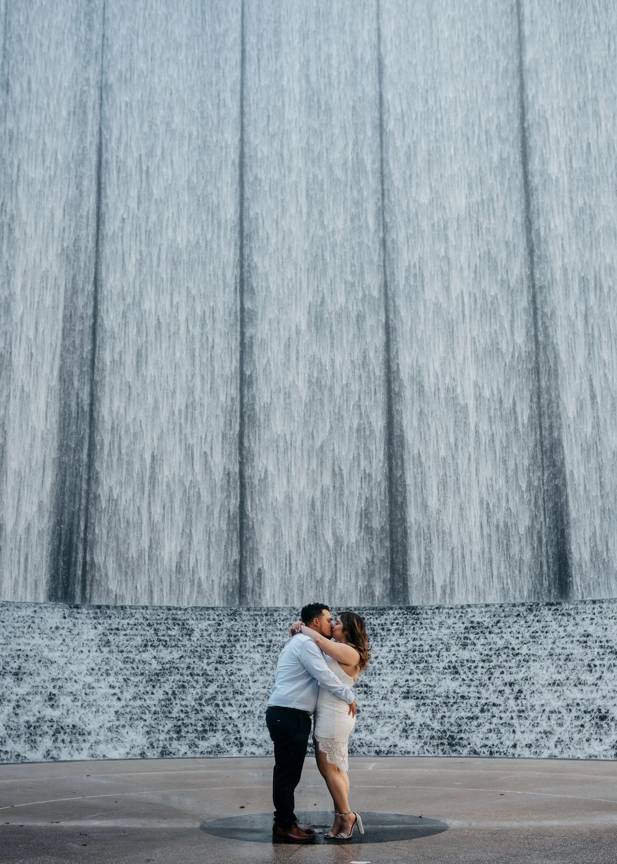 A couple embracing and kissing in front of a large waterfall with water cascading down the wall behind them.