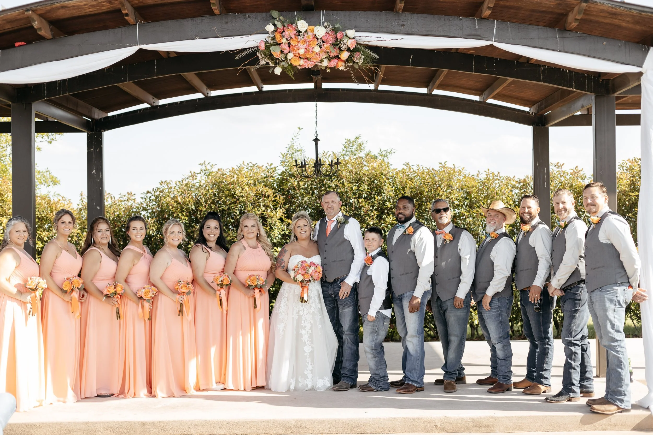 A wedding party on an outdoor stage with the bride and groom in the center, surrounded by bridesmaids in peach dresses on the left and groomsmen in vests and jeans on the right, under a decorated wooden arch.