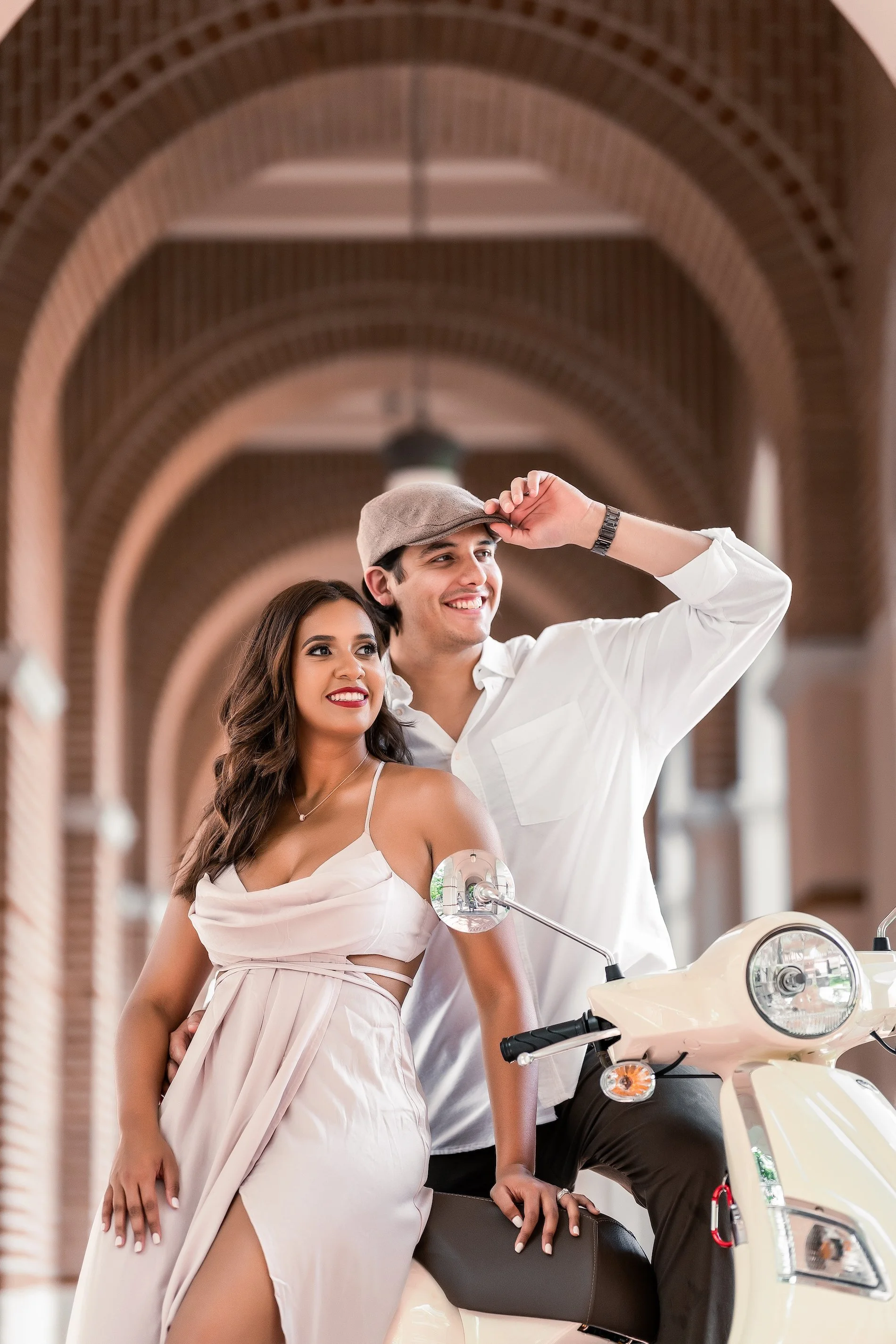 A smiling young couple with a scooter under arched brick bridges, woman in a cream dress and man in a white shirt and cap, in a bright, modern hallway.