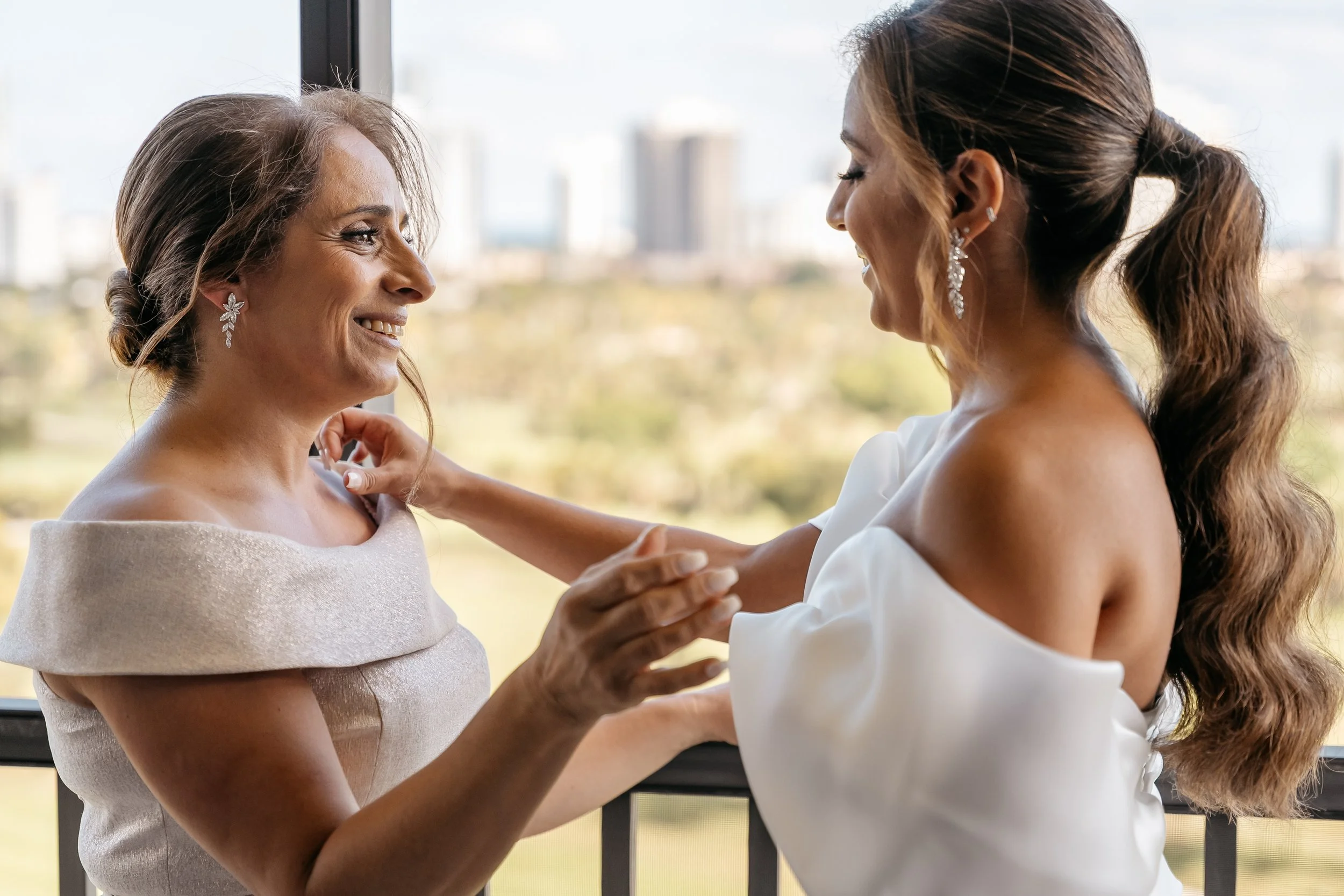 Two women, possibly a mother and daughter, smiling and holding hands near a window with a cityscape in the background.