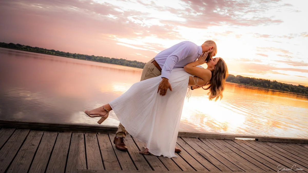 A couple kissing on a wooden dock by a lake at sunset, with the man dipping the woman backward.