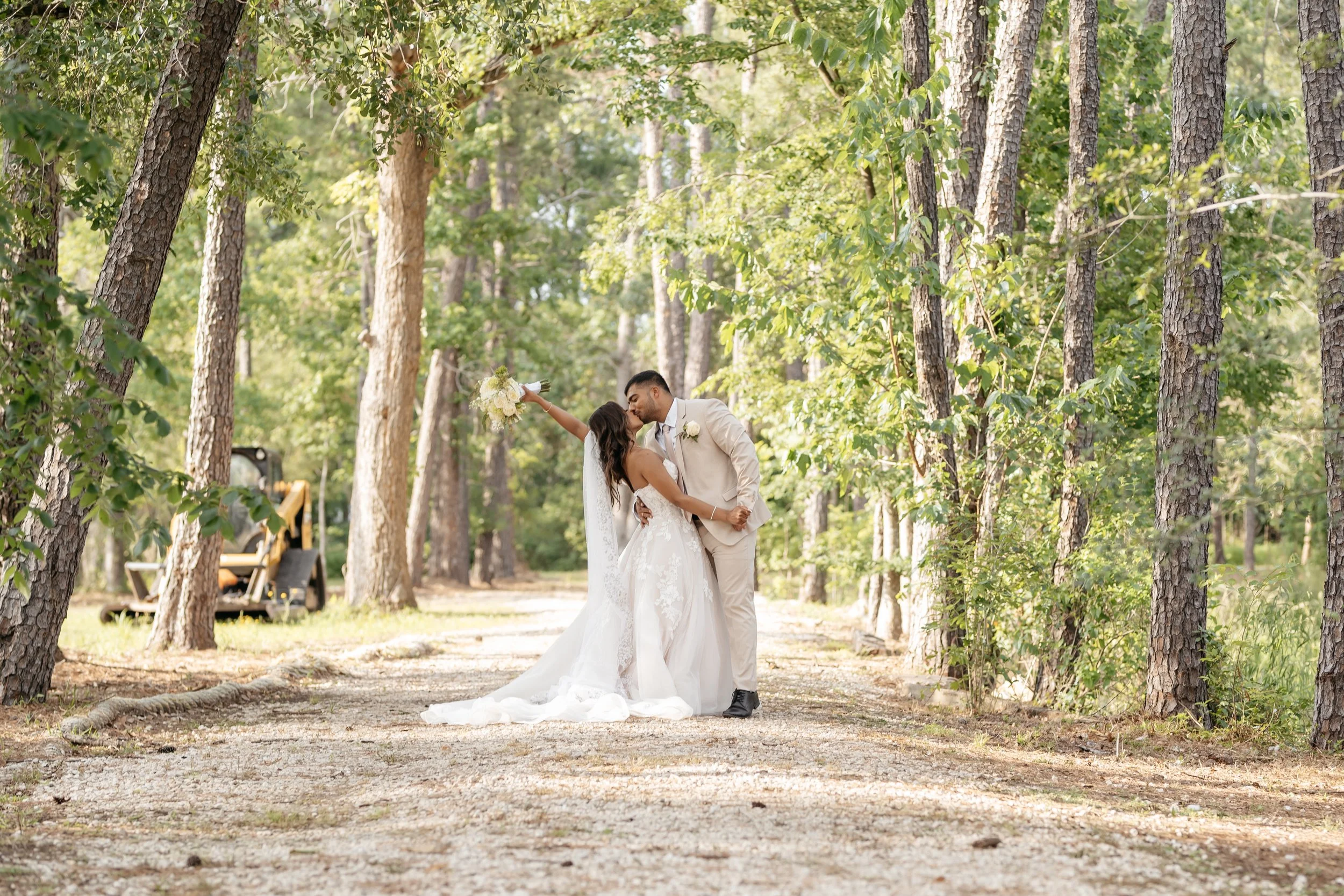 A bride and groom kissing in a forested area, with the bride holding a bouquet and the groom dressed in a light-colored suit.