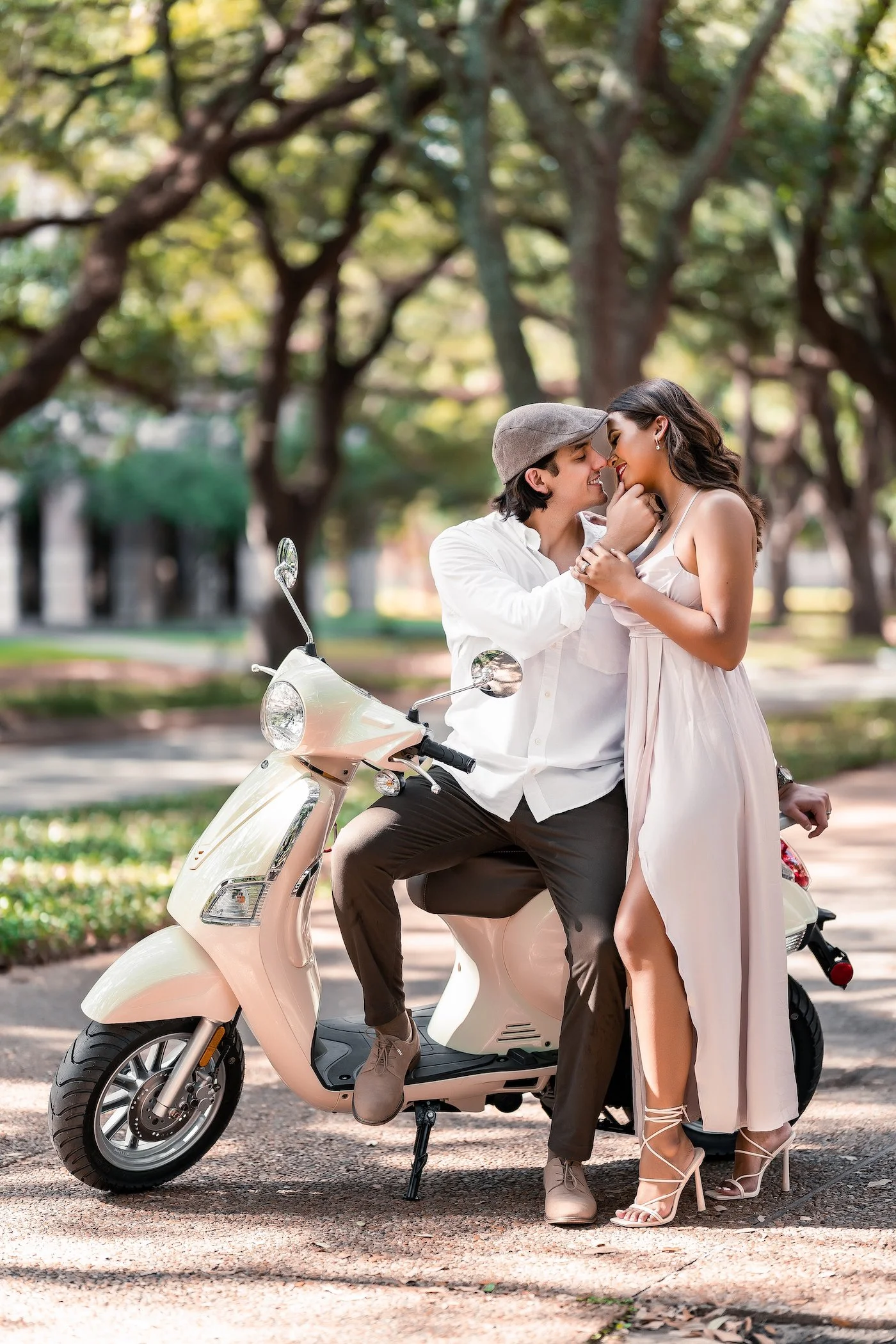 A romantic couple is on a scooter in a park, embracing and smiling at each other, with trees in the background.