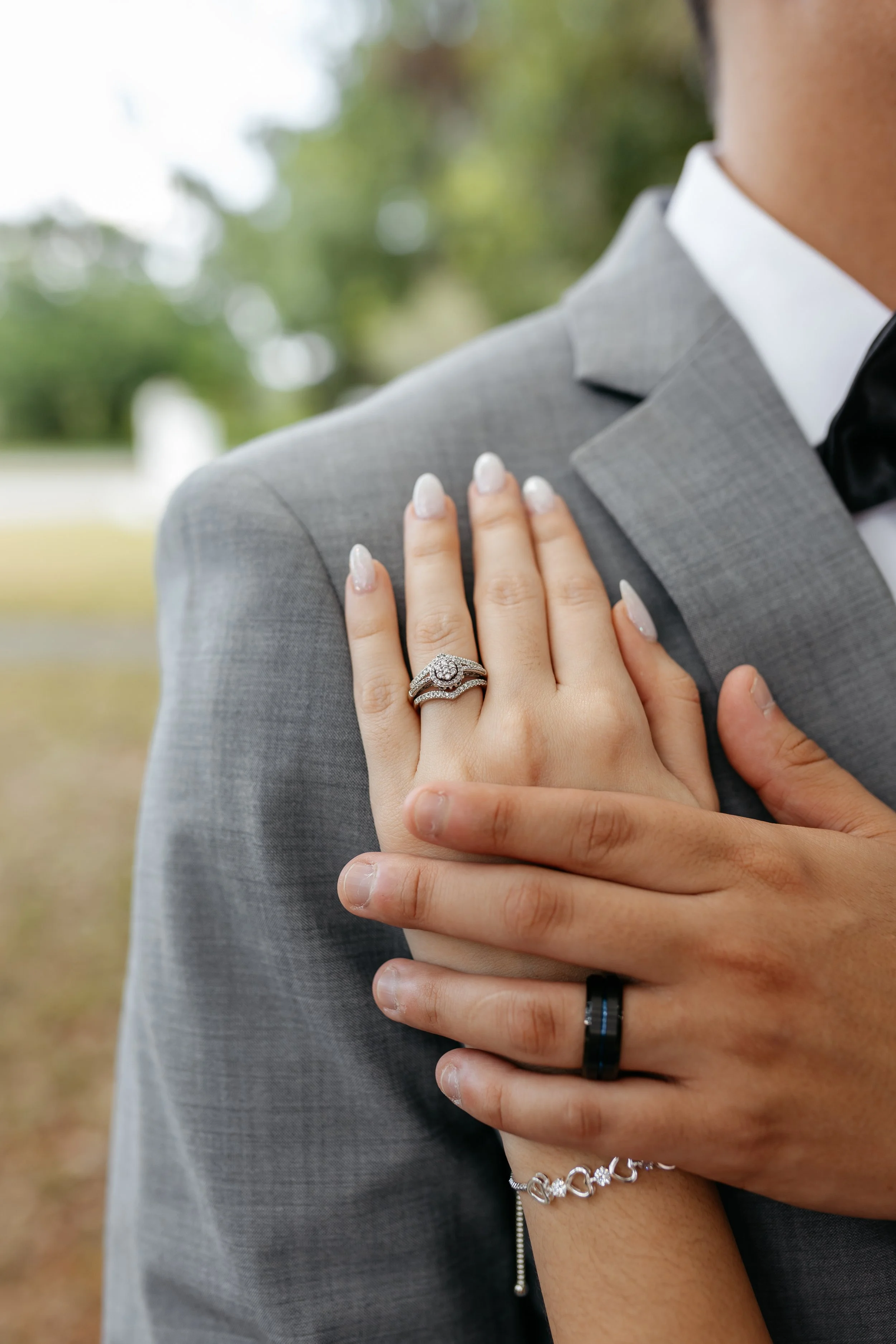 Close-up of a woman's hand with wedding rings resting on a man's shoulder, who is wearing a gray suit and black bow tie, outdoors with blurred greenery in the background.