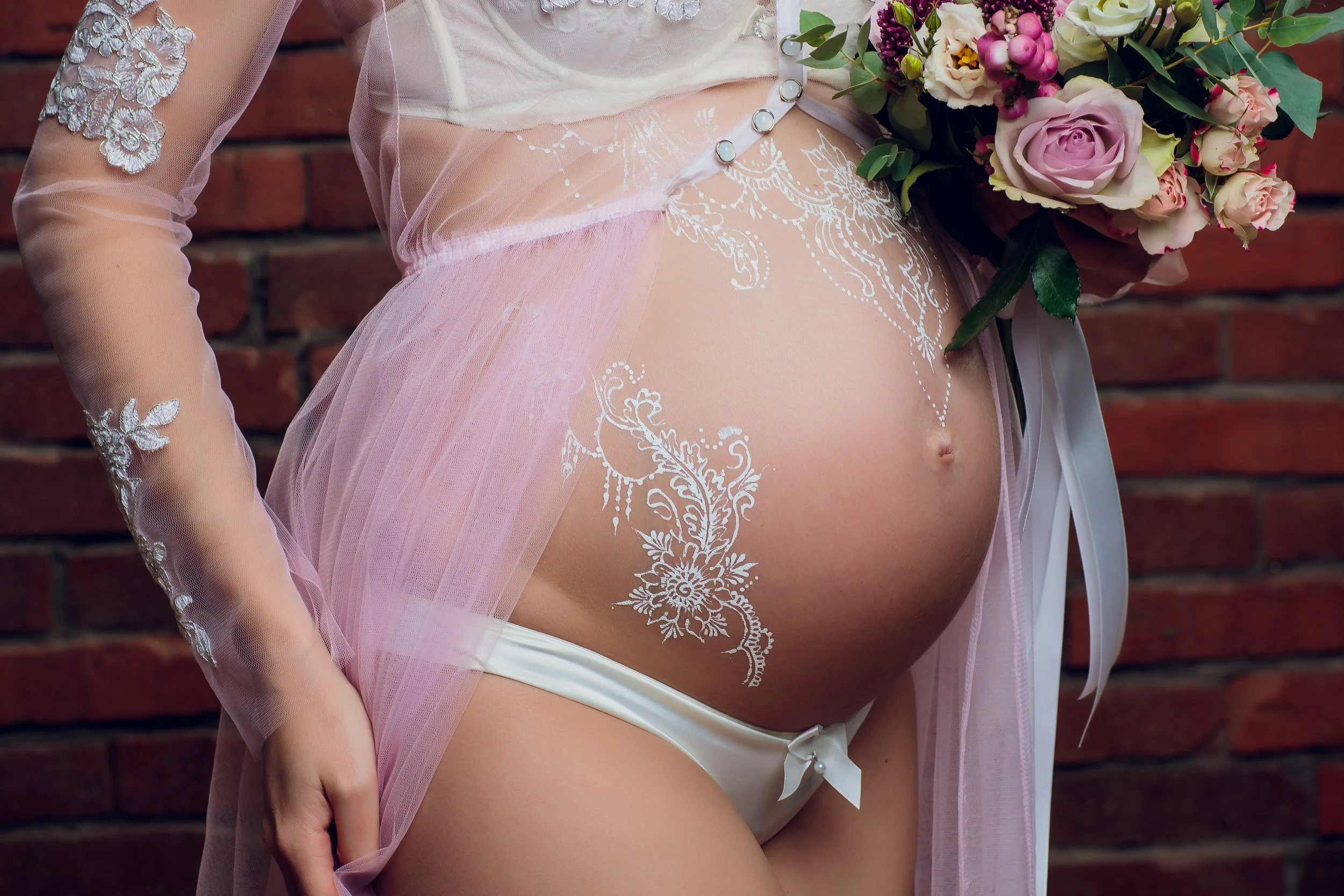 Close-up of a pregnant woman in sheer pink lace and tulle lingerie, holding a bouquet of flowers, standing against a brick wall.