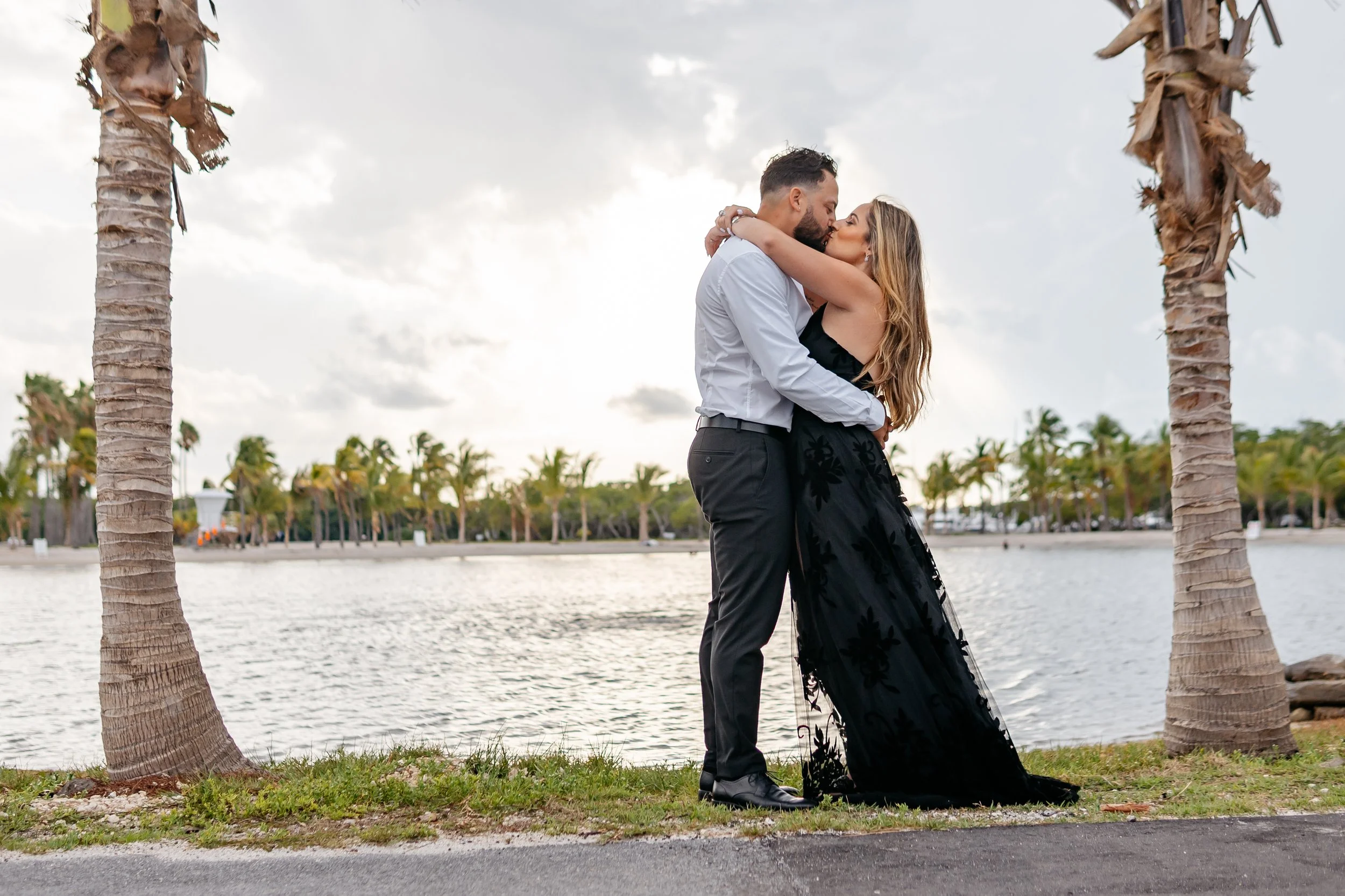 A couple in formal attire embracing and kissing by a waterfront with palm trees in the background.