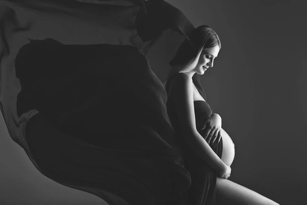 Black and white photo of a pregnant woman sitting and smiling, with a woman holding a large piece of fabric behind her.