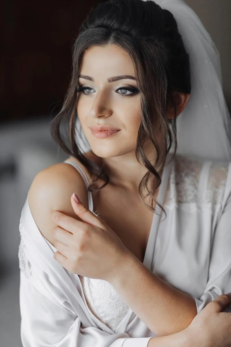 Bride in a white lace robe and veil with soft curls and bridal makeup, photographed during wedding preparations by a luxury wedding photographer
