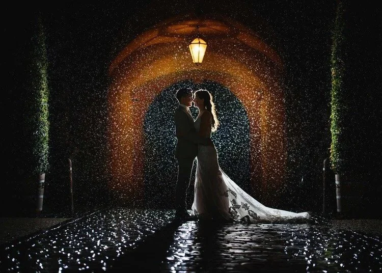 A couple in wedding attire, standing close and embracing under a streetlamp, in the rain with water droplets falling around them at night.