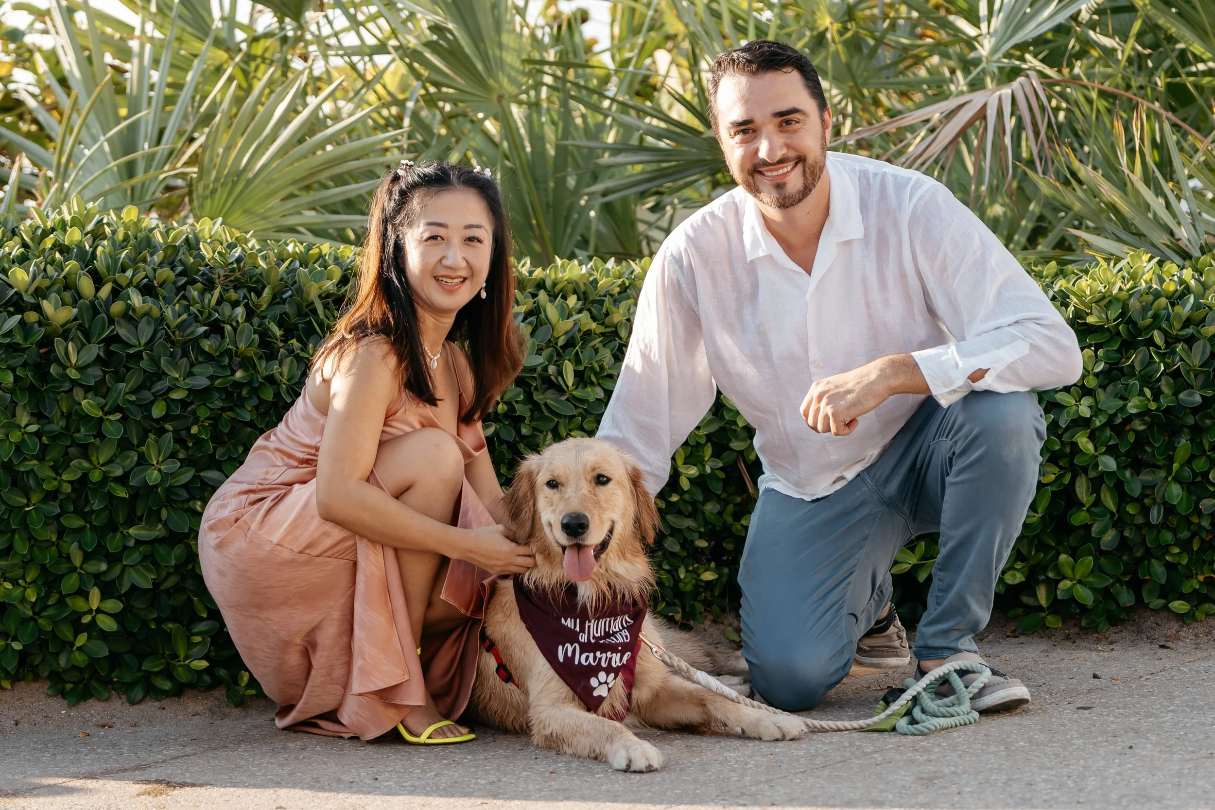 A woman in a pink satin dress and a man in a white shirt kneel next to a golden retriever wearing a maroon bandana that reads, 'All Humans Give Love, Marrie.' The woman is petting the dog, and the man has his hand on the dog's head. They are outdoors
