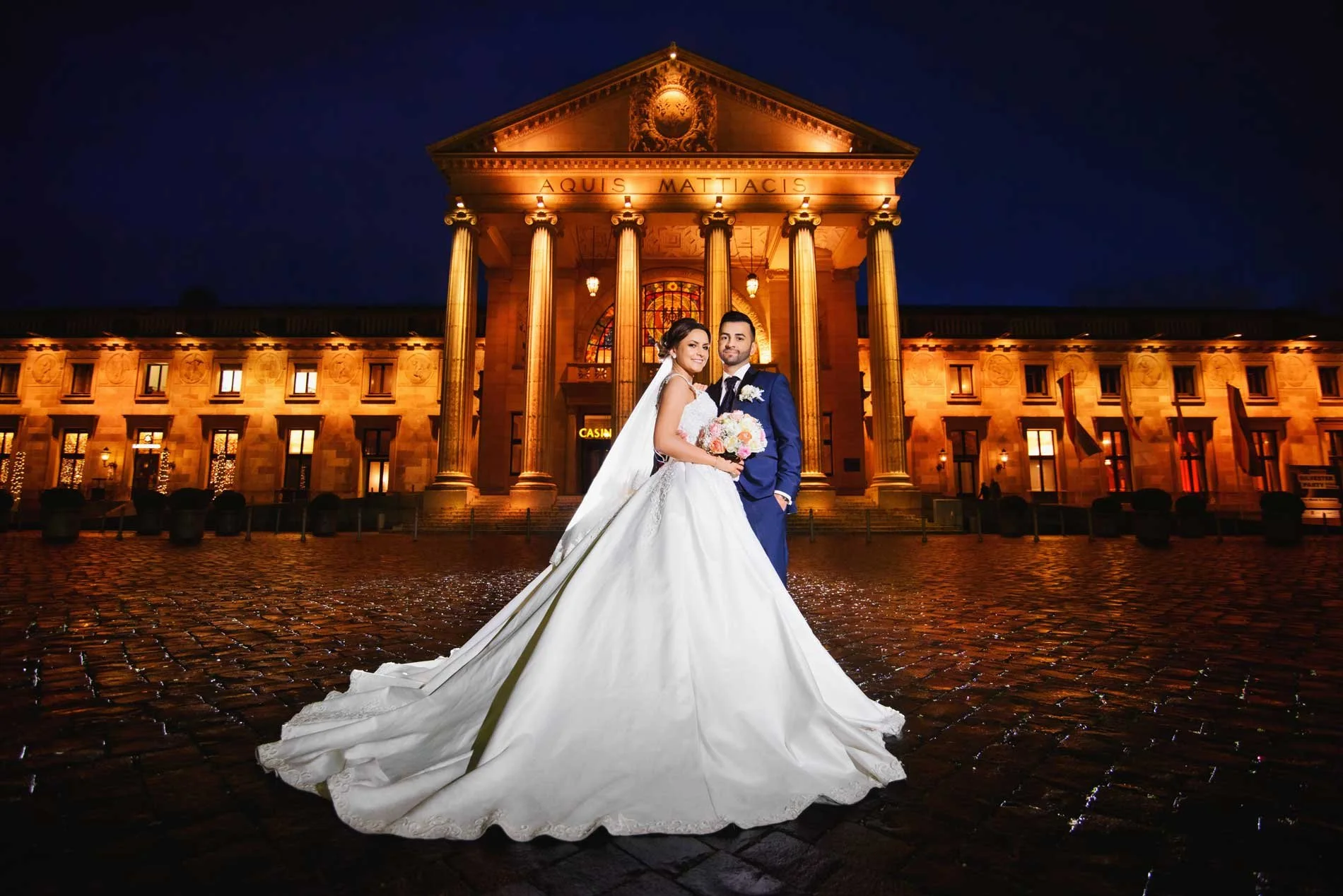 A bride and groom in wedding attire standing in front of a large illuminated building with columns at night, with cobblestone ground.