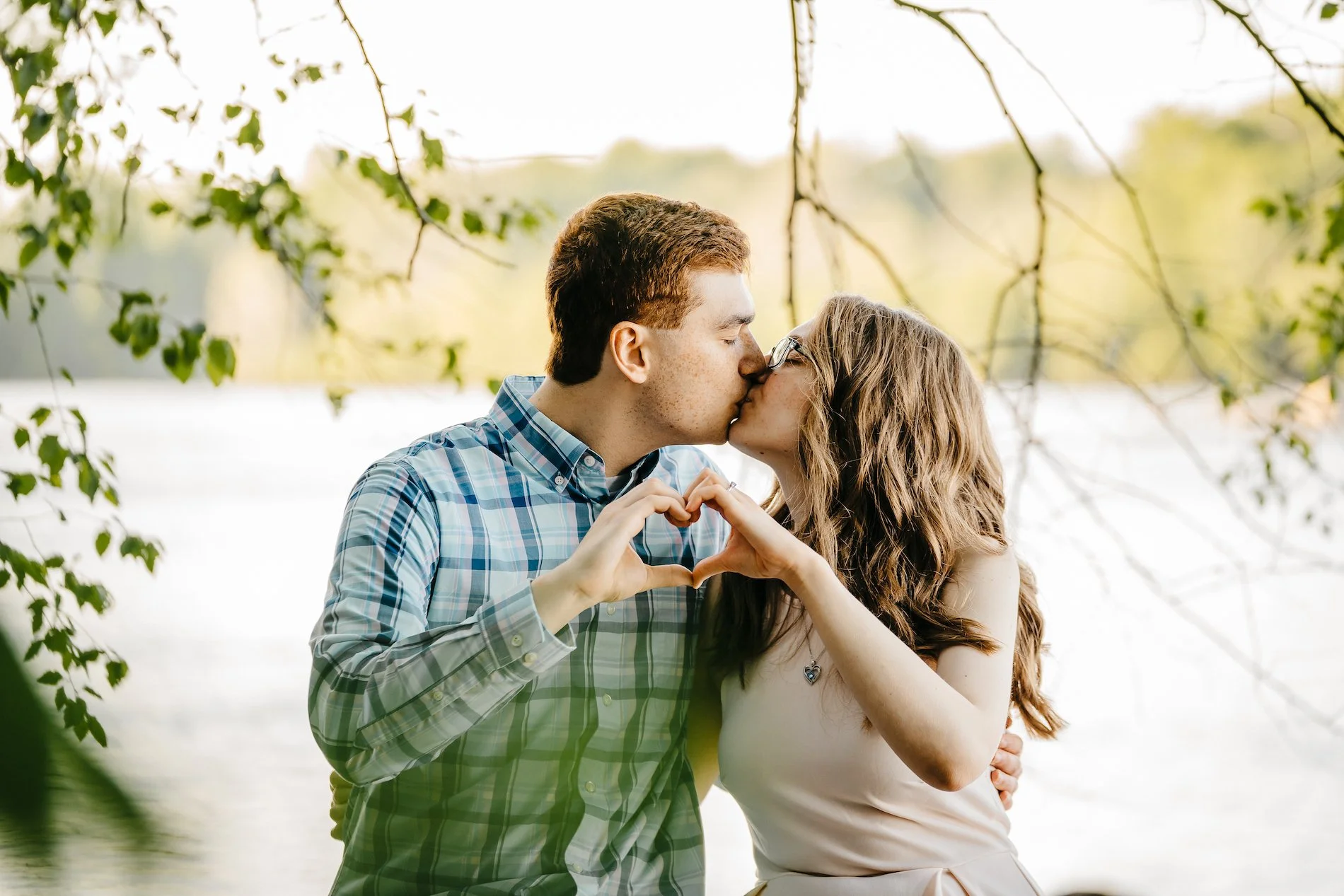 A young couple sharing a kiss outdoors near a lake, making a heart shape with their hands.
