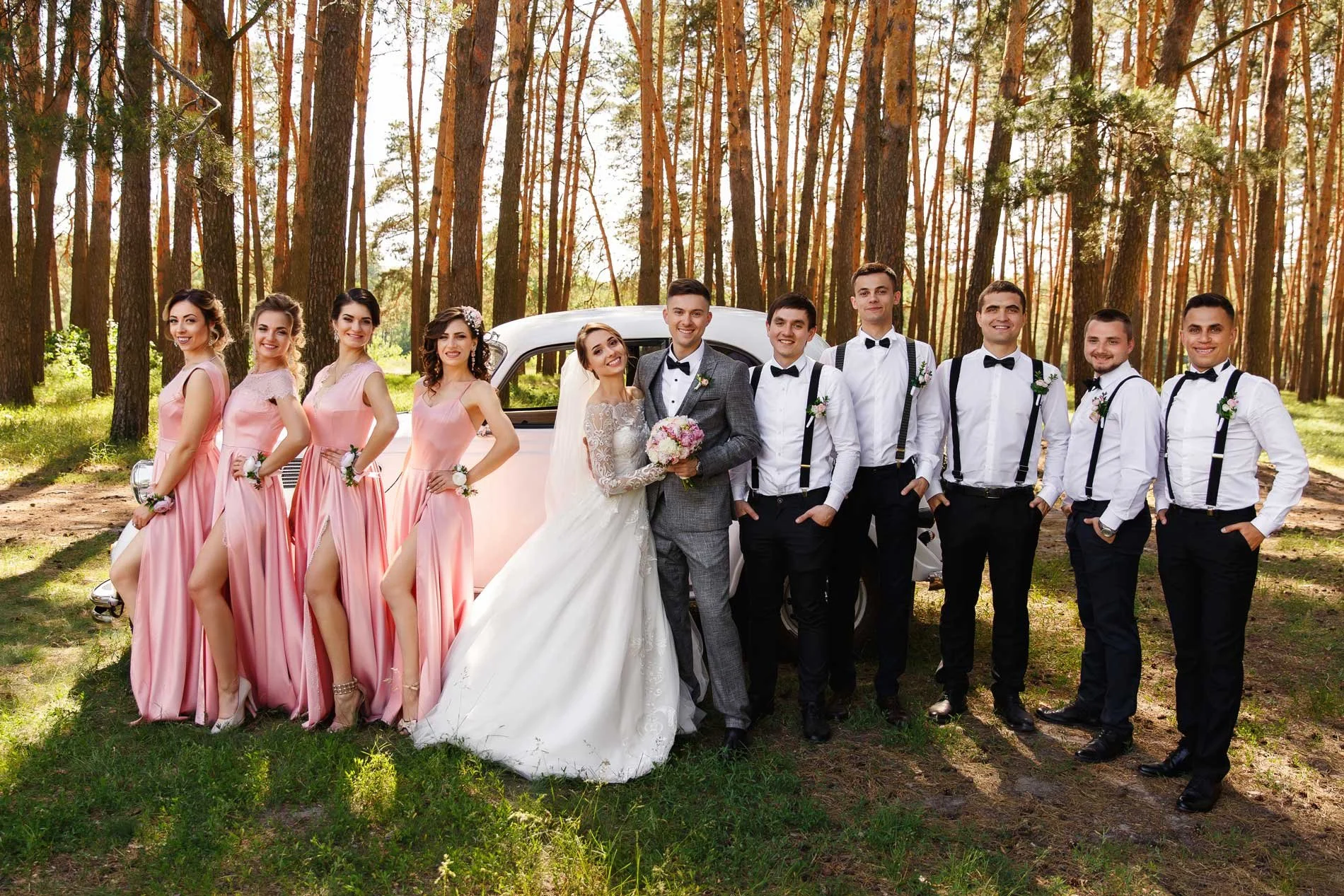 A wedding party standing outdoors in a forest, with the bride and groom in the center, surrounded by bridesmaids in pink dresses on the left and groomsmen in white shirts with black suspenders on the right, all smiling.