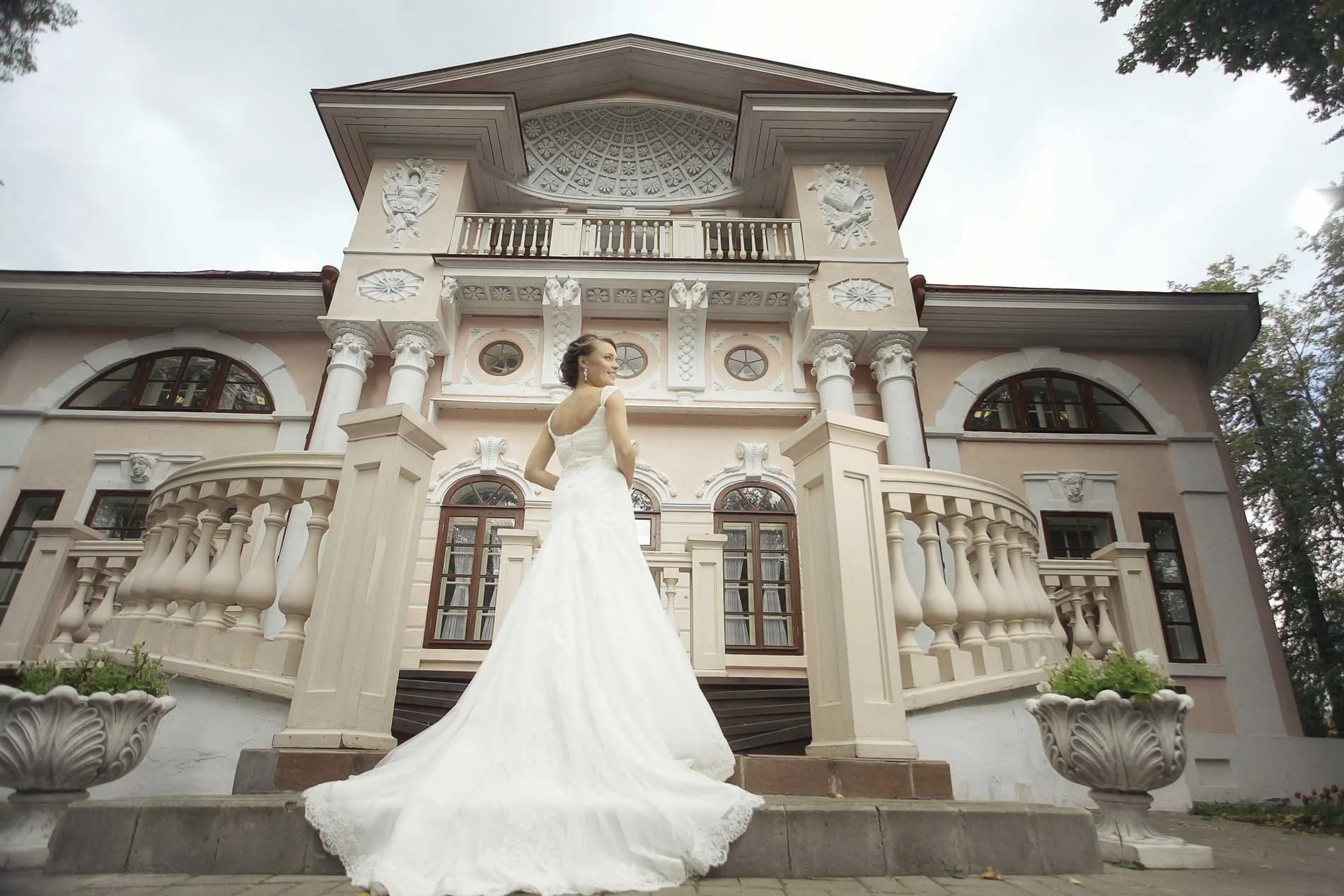 A woman in a white wedding dress standing on stairs in front of a large ornate house with columns and arched windows.