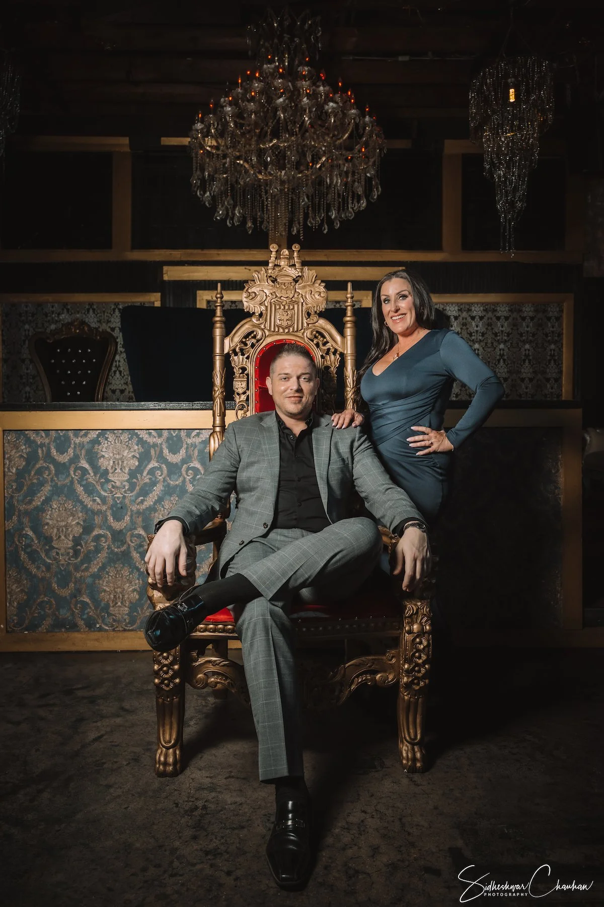 A man in a checkered gray suit and black shoes sitting on an ornate red velvet and gold throne, with a woman in a dark teal dress standing beside him, her hand resting on his shoulder, inside a dimly lit room with elaborate chandeliers and classic de