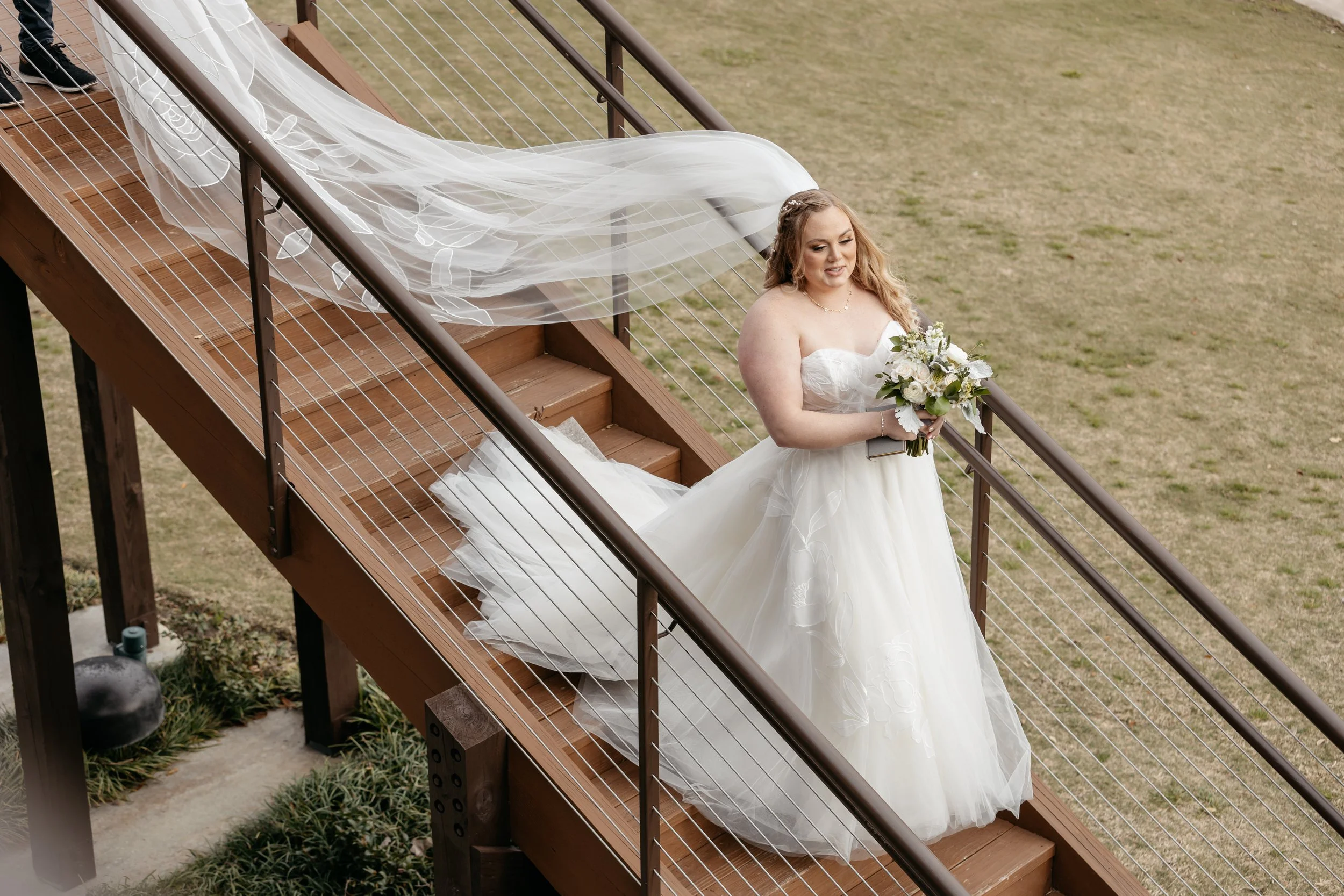 A bride in a strapless white wedding gown holding a bouquet of flowers standing on a wooden staircase outdoors.