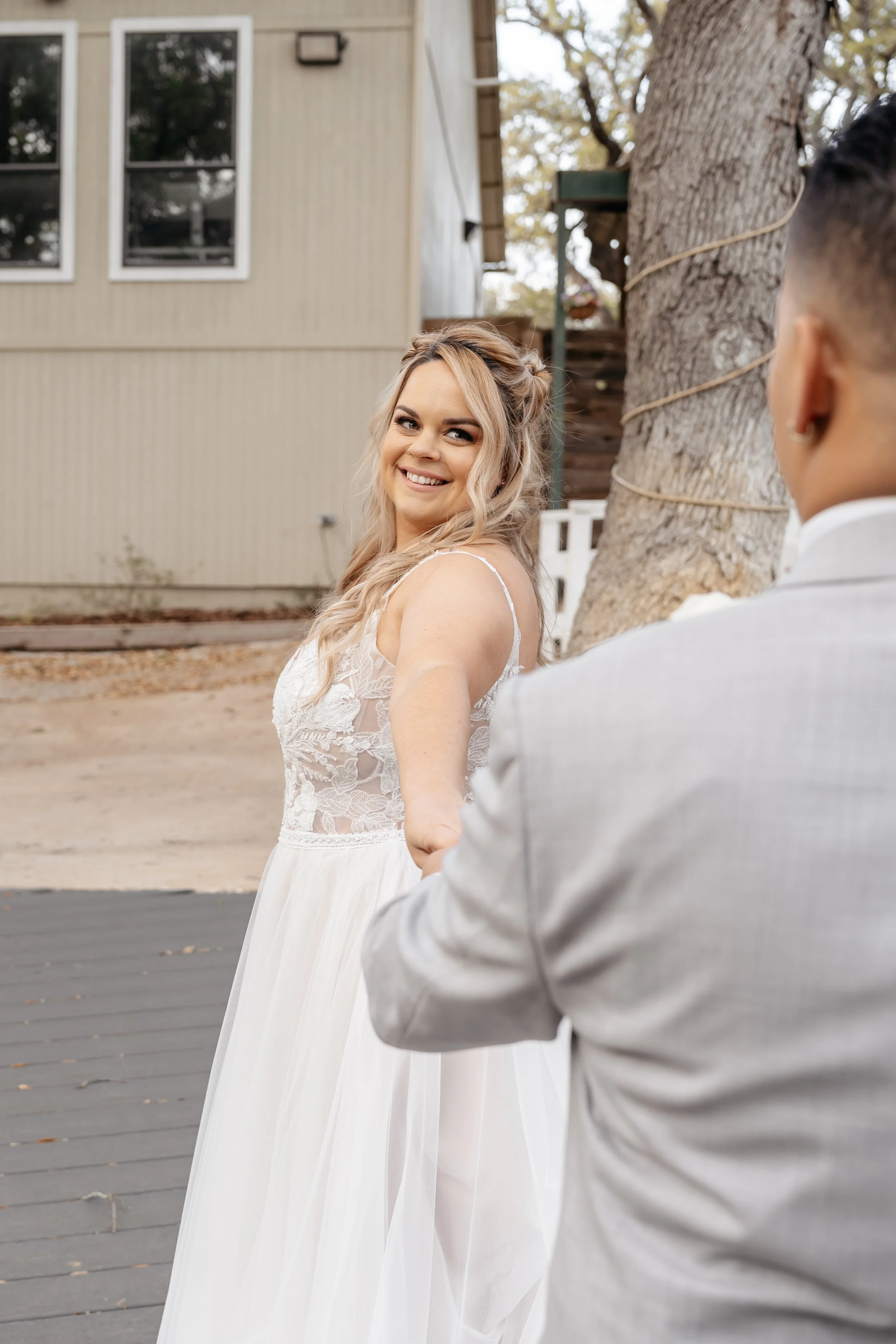 A bride smiling happily at her groom outdoors, holding his hand, in front of a tree and a house.