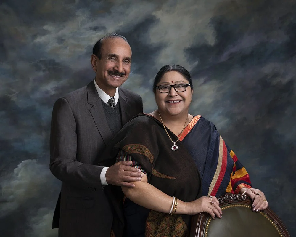 A middle-aged Indian couple, man with a mustache in a dark suit and tie, woman wearing glasses and a traditional colorful sari, standing against a dark cloudy background.