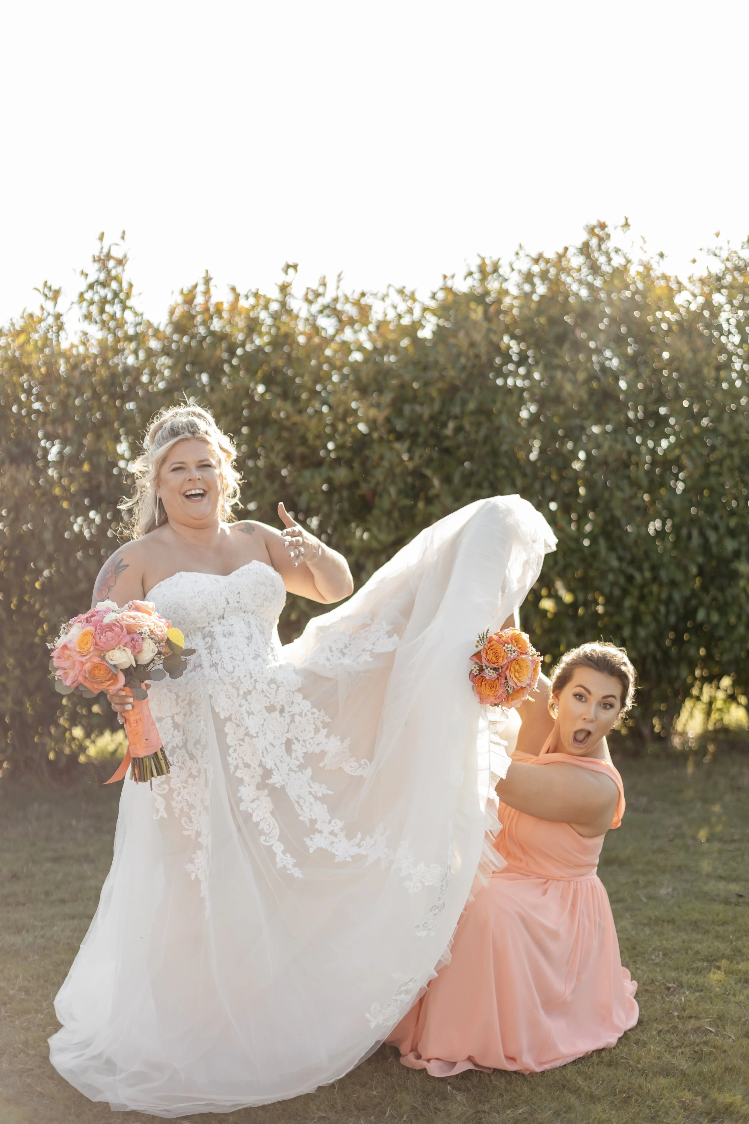 Bride in a white wedding dress holding a bouquet of pink and peach roses, being playfully lifted by her bridesmaid in a peach dress, both smiling with a background of green foliage.