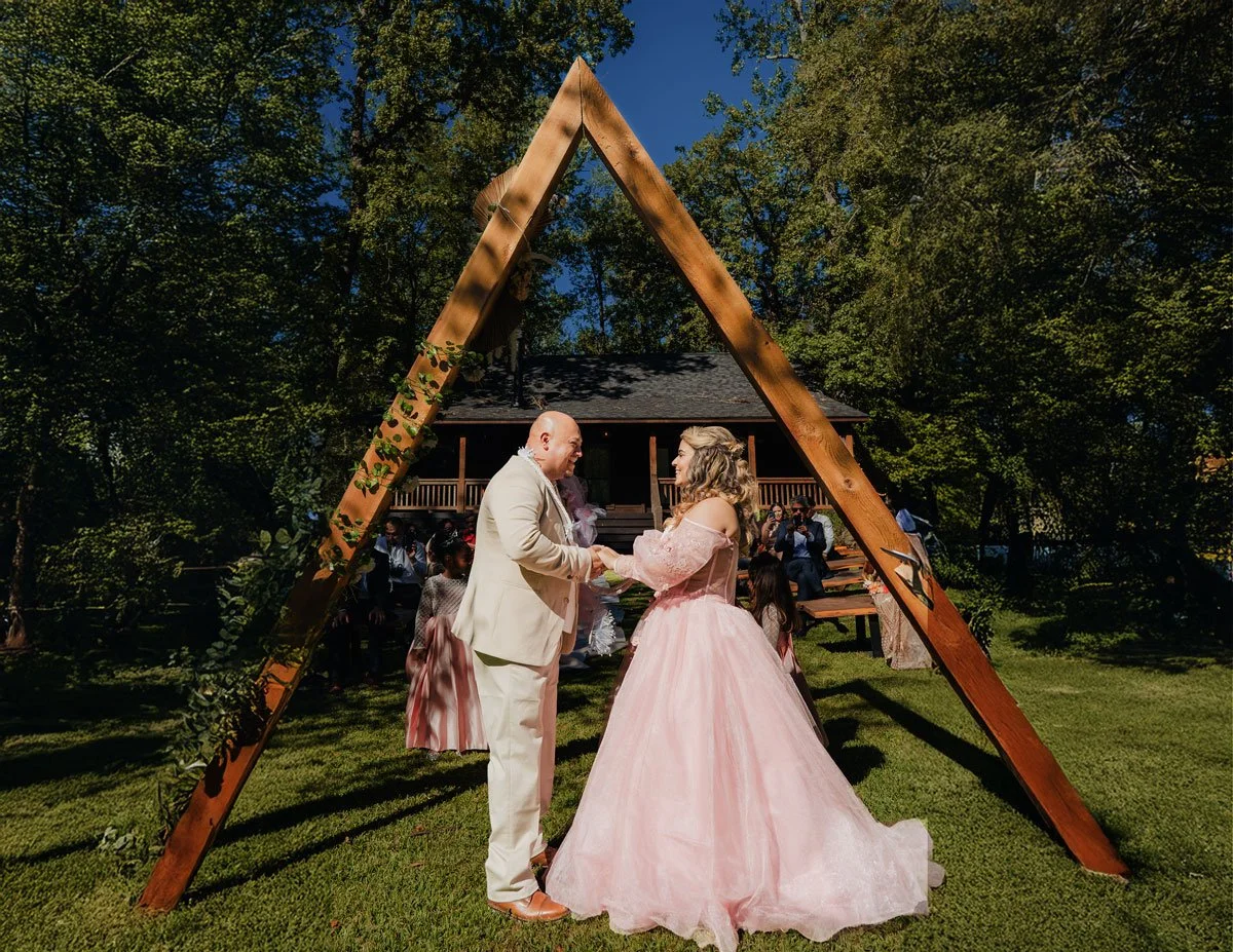 A couple getting married outdoors beneath a wooden A-frame arch decorated with green foliage, with guests seated behind them on a grassy lawn and a house in the background.