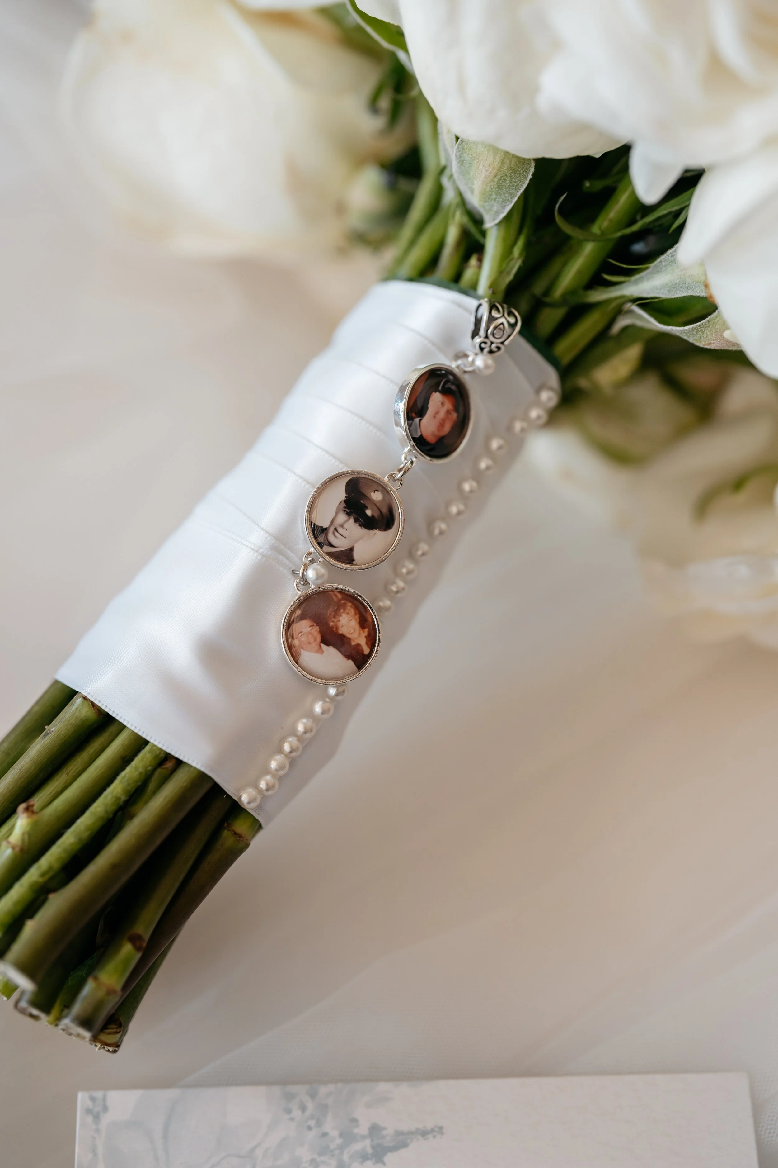 A bridal bouquet with white flowers wrapped with white satin ribbon and decorated with a photo keepsake charm containing three black-and-white and color photos of people, resting on a white surface.