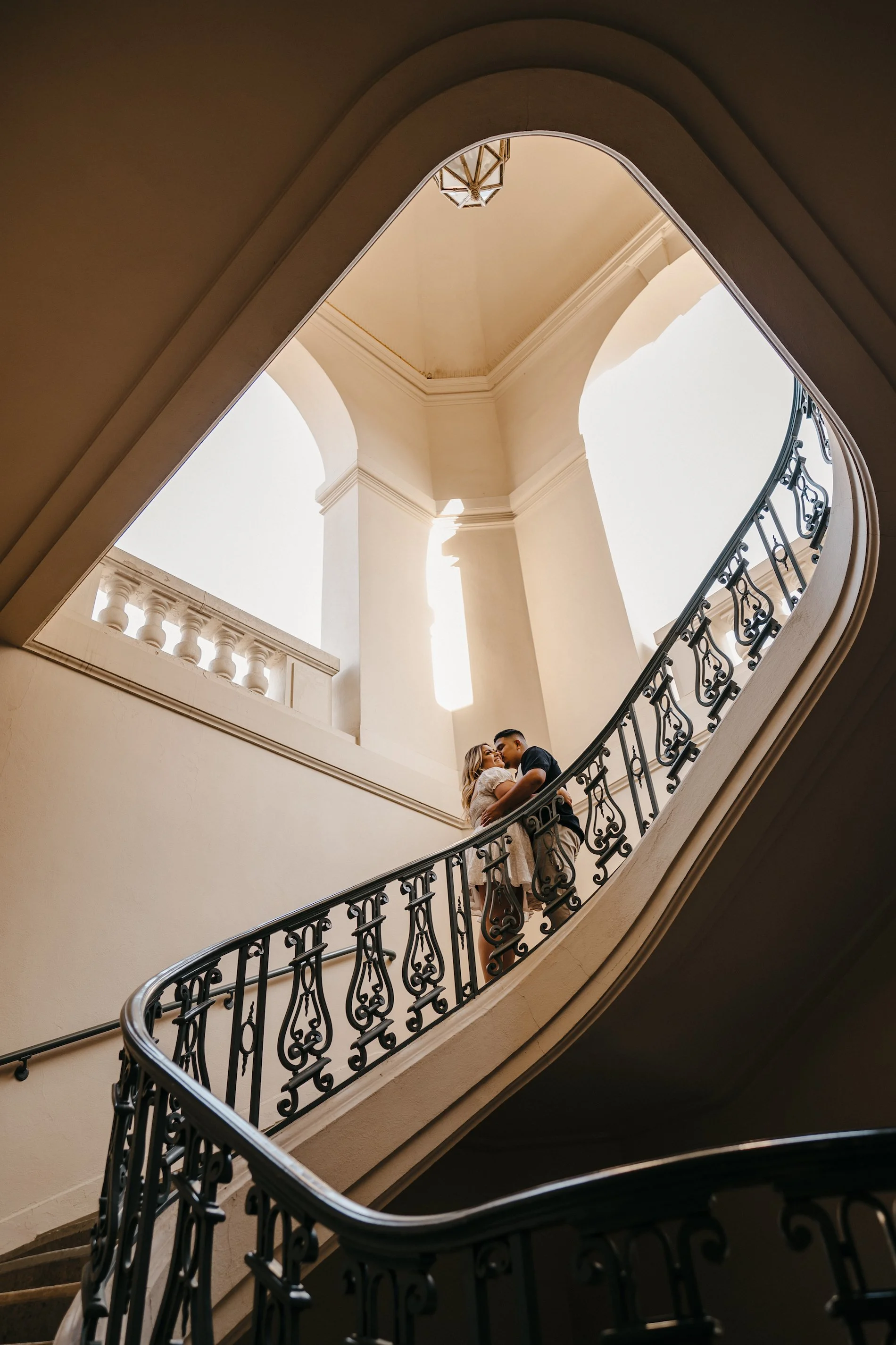 A couple standing on a spiral staircase, captured from below, with natural light and architectural details in the background.